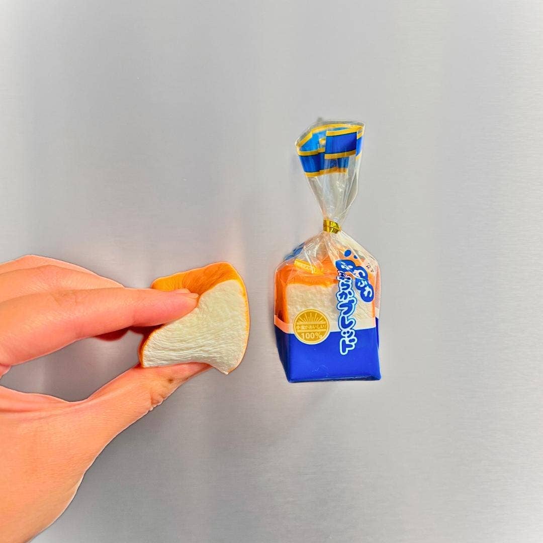 A hand holds a miniature toy slice of bread next to a miniature toy loaf of bread packaged in a blue, white, and yellow bag, set against a plain gray background.