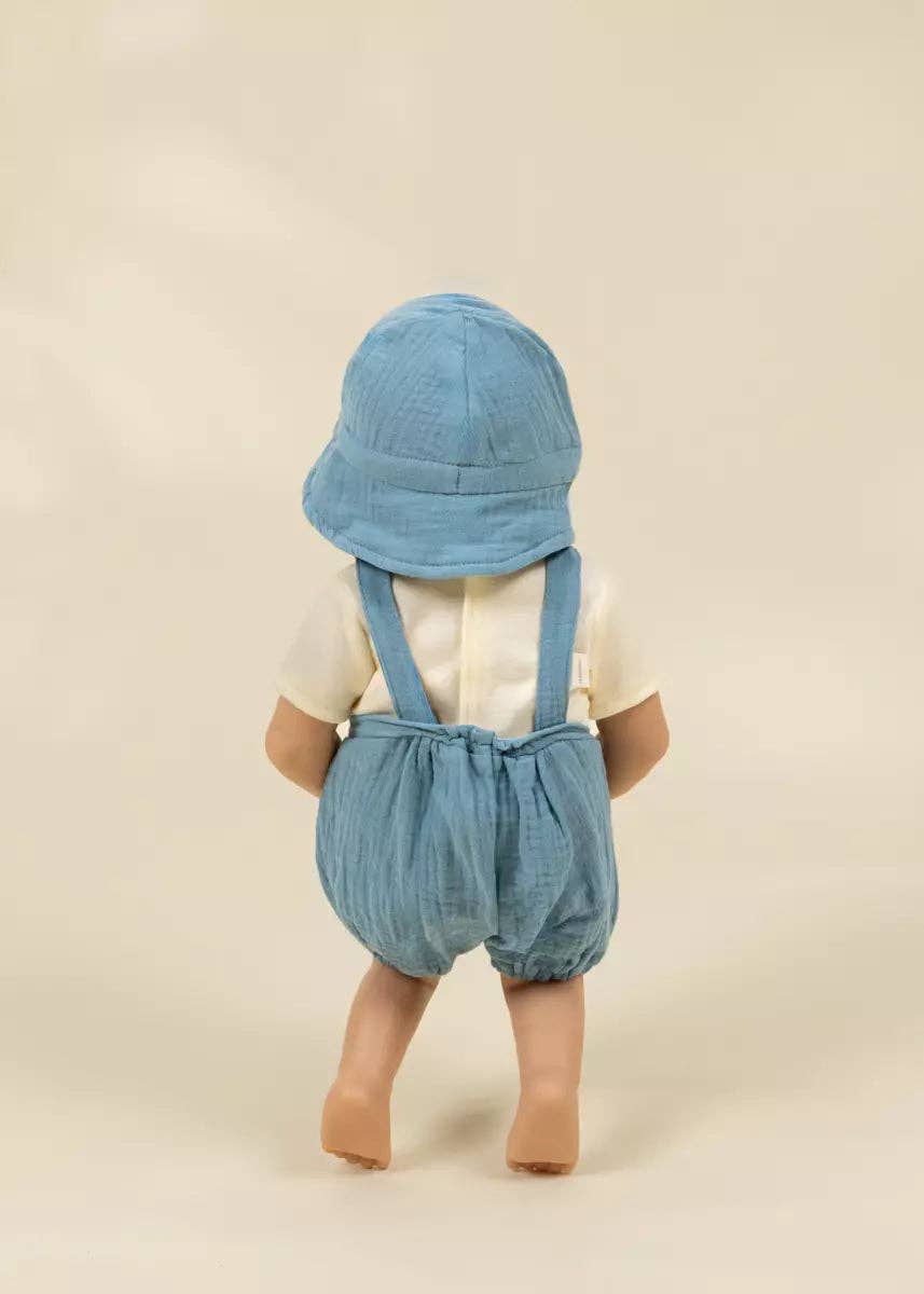 A baby doll viewed from the back, wearing a light blue sun hat, blue denim-style suspenders, and a cream short-sleeve top, standing against a plain beige studio background.