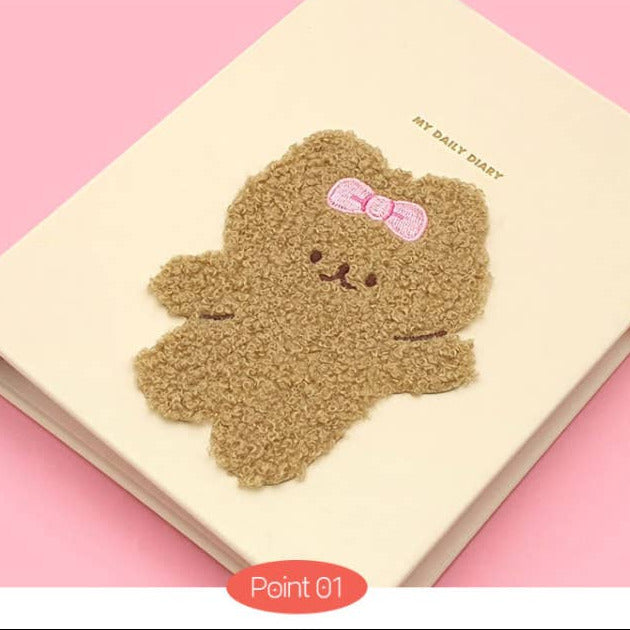 A cream-colored daily diary with a textured brown teddy bear applique wearing a pink bow, set against a solid pink background.