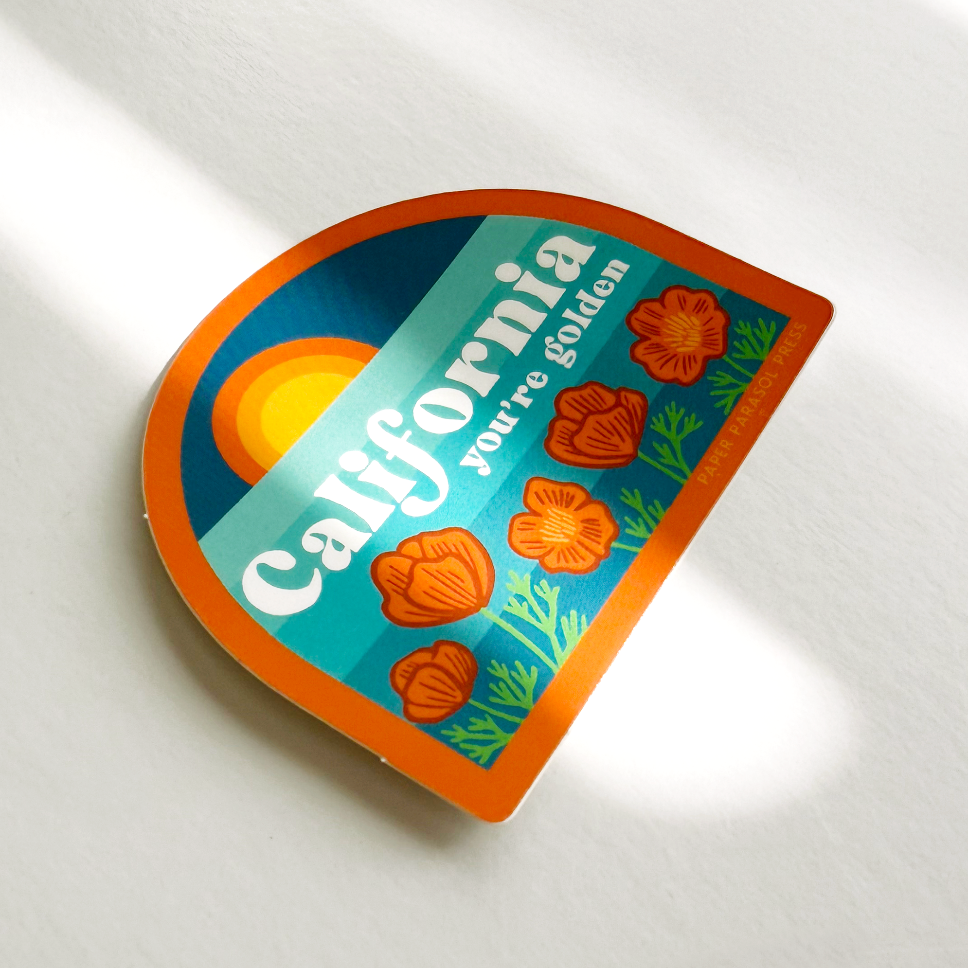 A die-cut sticker with a retro California design, featuring orange poppies, a yellow sun, and the text 'California you're golden', placed on a white textured surface with soft diagonal shadows.