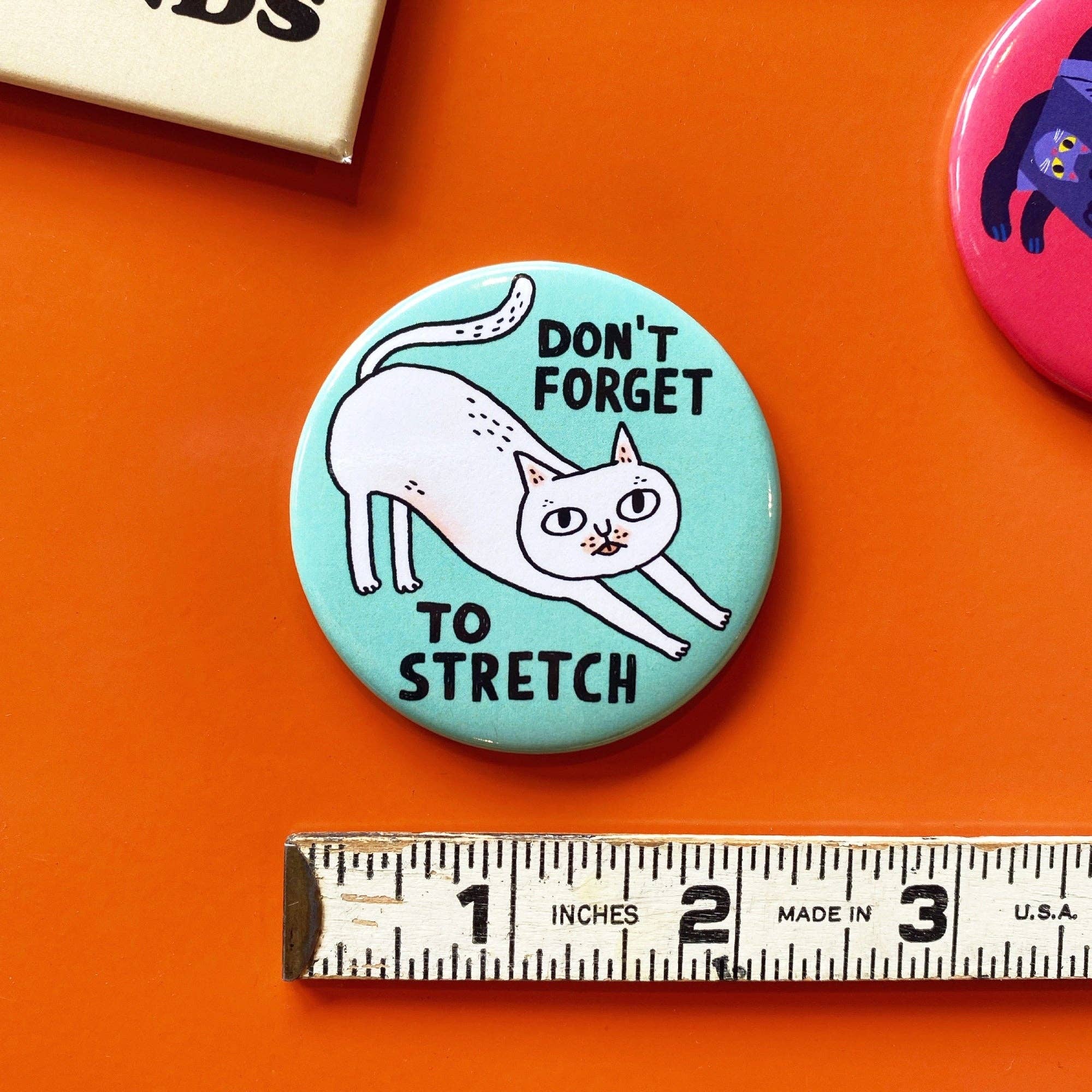 Button badge with a cat illustration and text 'Don't forget to stretch' on an orange background, with a ruler for scale.