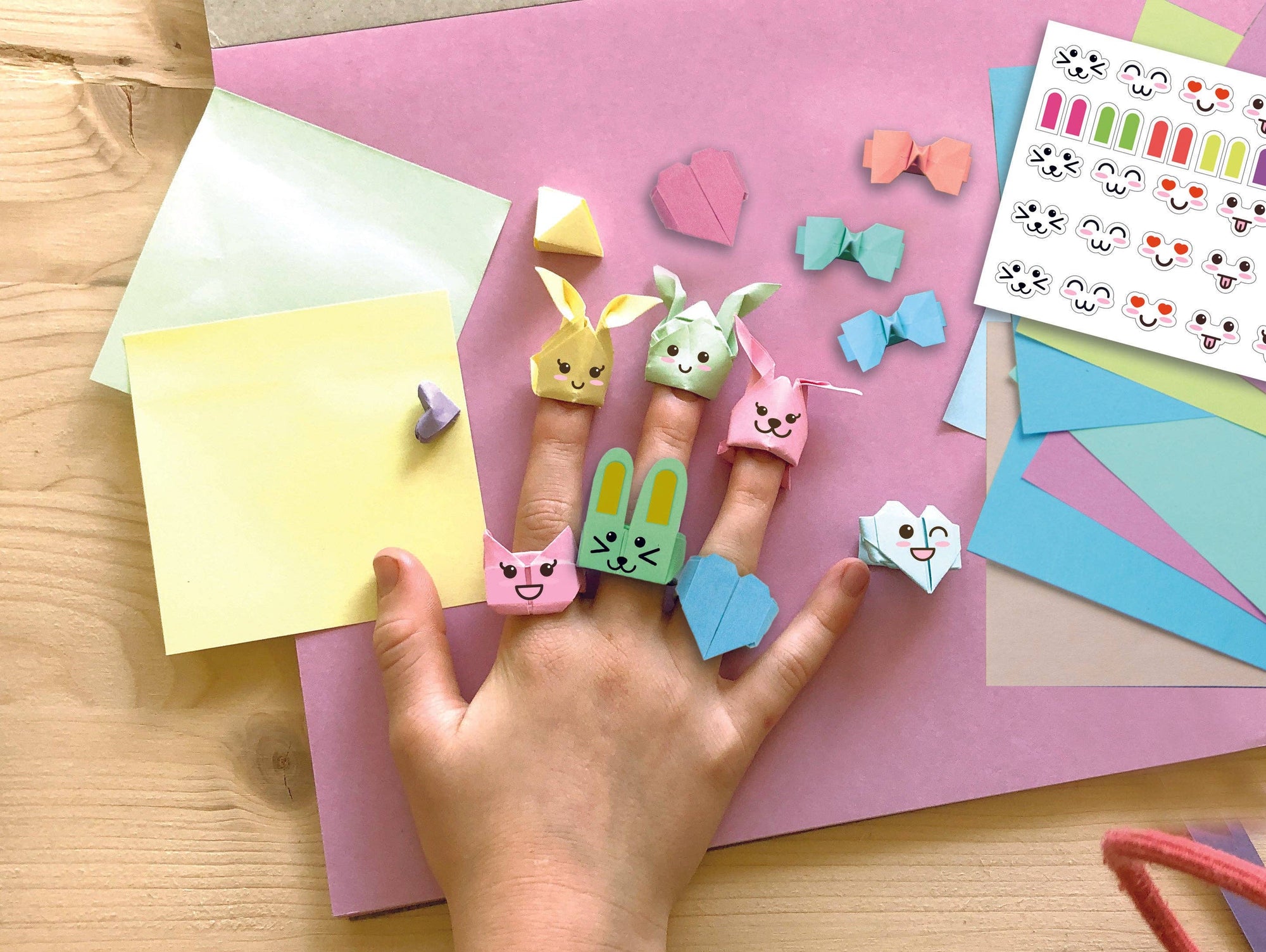 Finger puppets on a child's hand with colorful paper and stickers on a pink surface