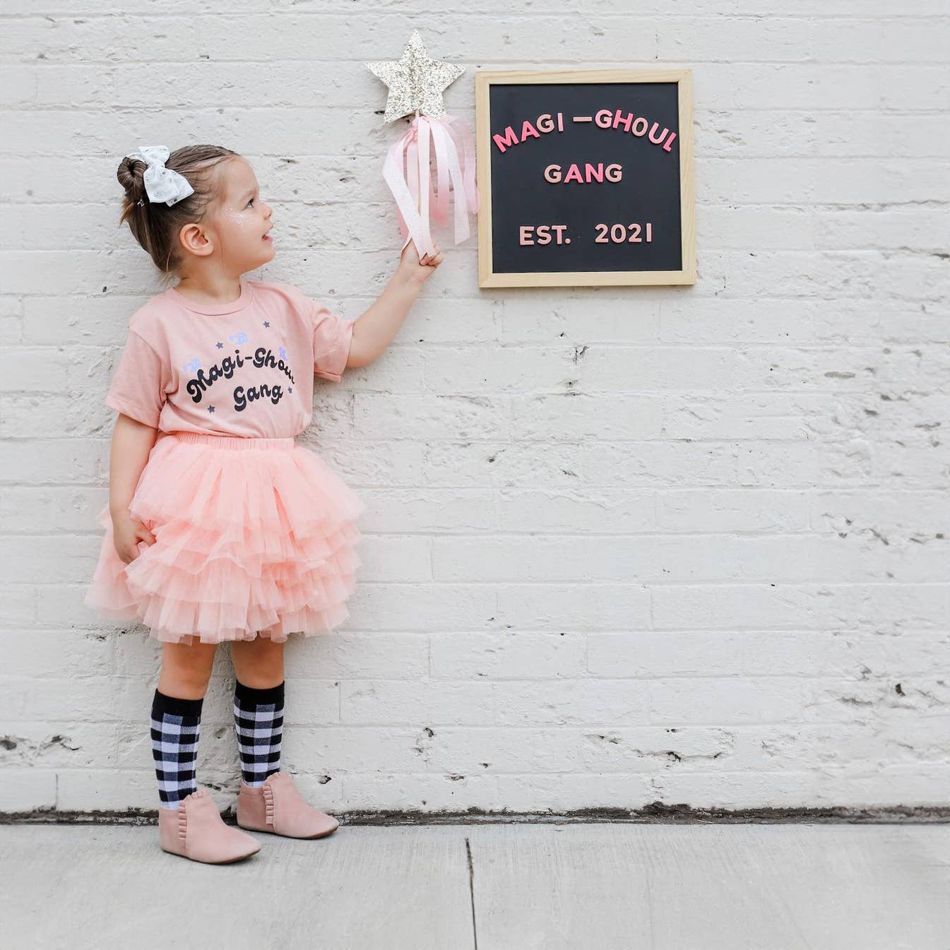 Child in pink outfit with 'Magi-Ghoul Gang' shirt and tutu, standing next to a chalkboard sign against a white brick wall.