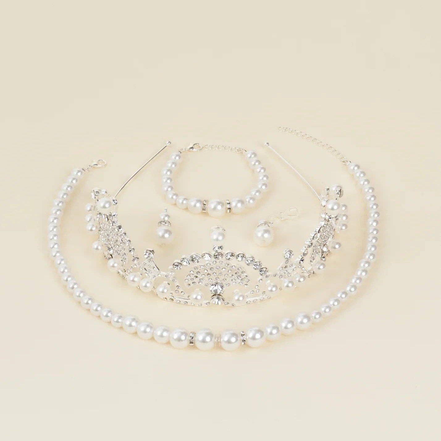 A silver and pearl bridal jewelry set featuring a tiara, collar necklace, bracelet, and earrings, arranged on a solid cream background.