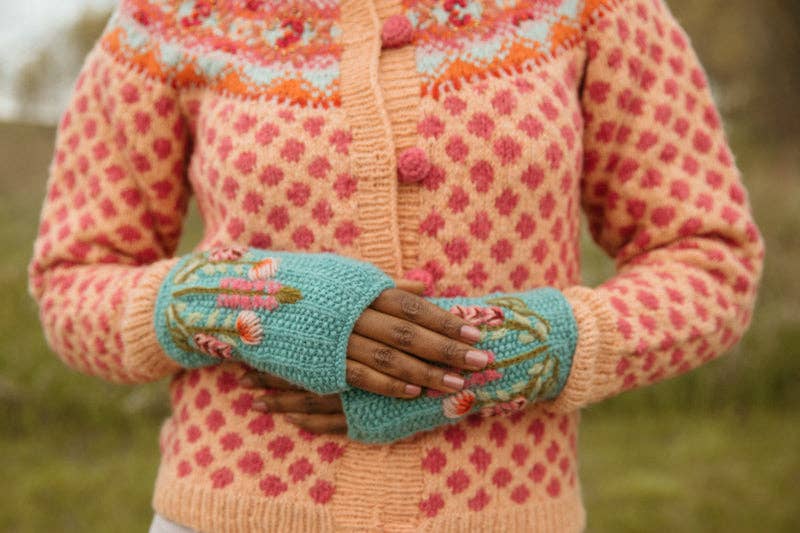 A person wearing a peach knit cardigan with pink floral patterns and turquoise fingerless gloves with floral embroidery, standing outdoors against a blurred green background.