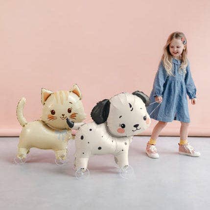 A young girl in a blue dress stands next to two large foil balloons shaped like a cream-colored cat and a white Dalmatian puppy, set against a soft pink studio background.