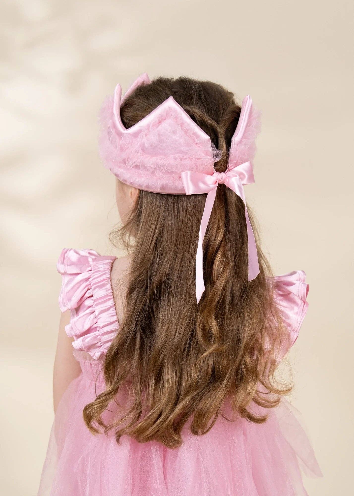 A young girl viewed from behind wearing a pink crown headband with a ribbon bow and a pink tulle dress against a soft beige background.