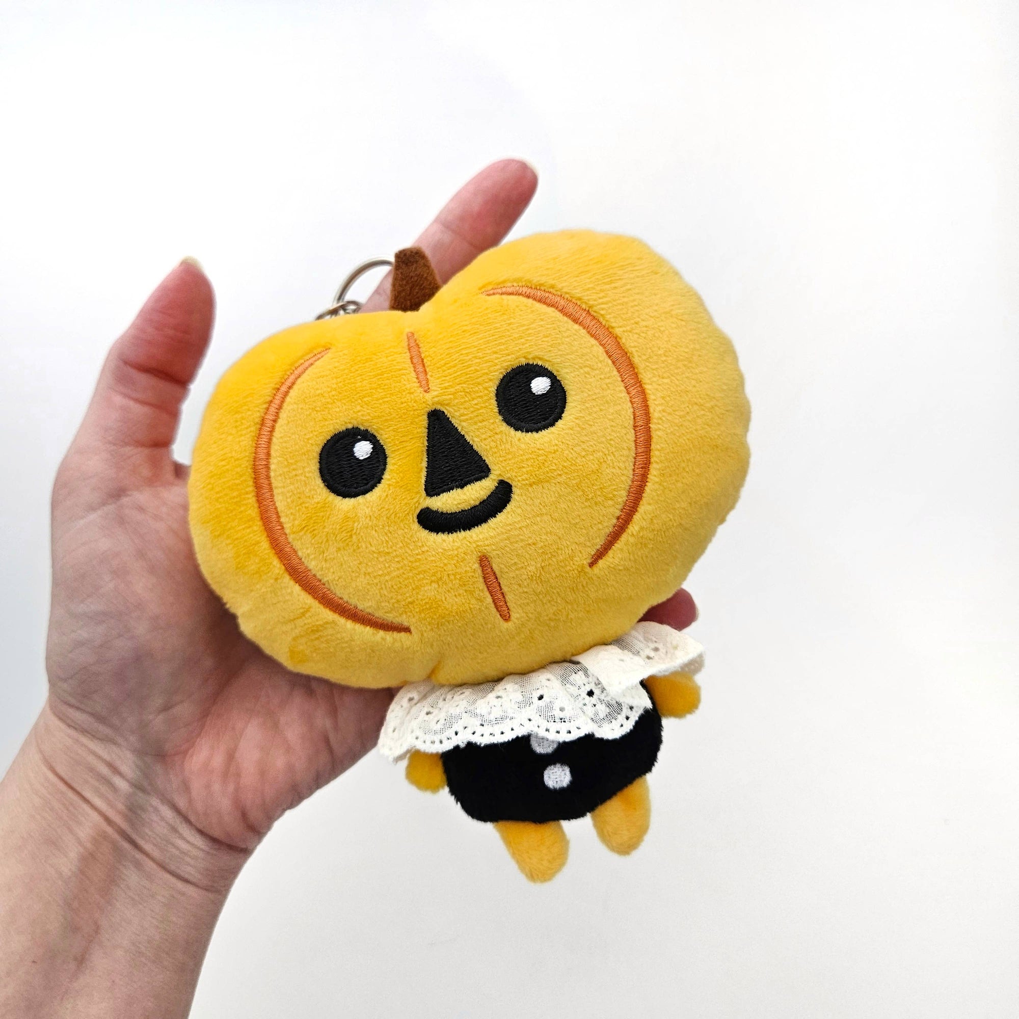 Plush keychain shaped like a orange punpkin character with a black nose and white lace trim on a white background