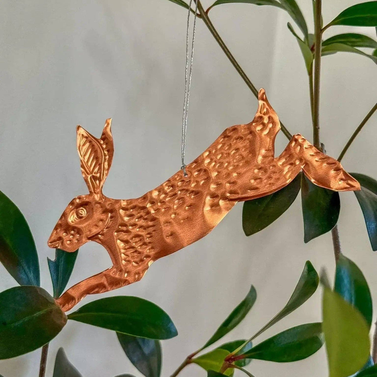 Copper hare ornament hanging from a branch with green leaves.
