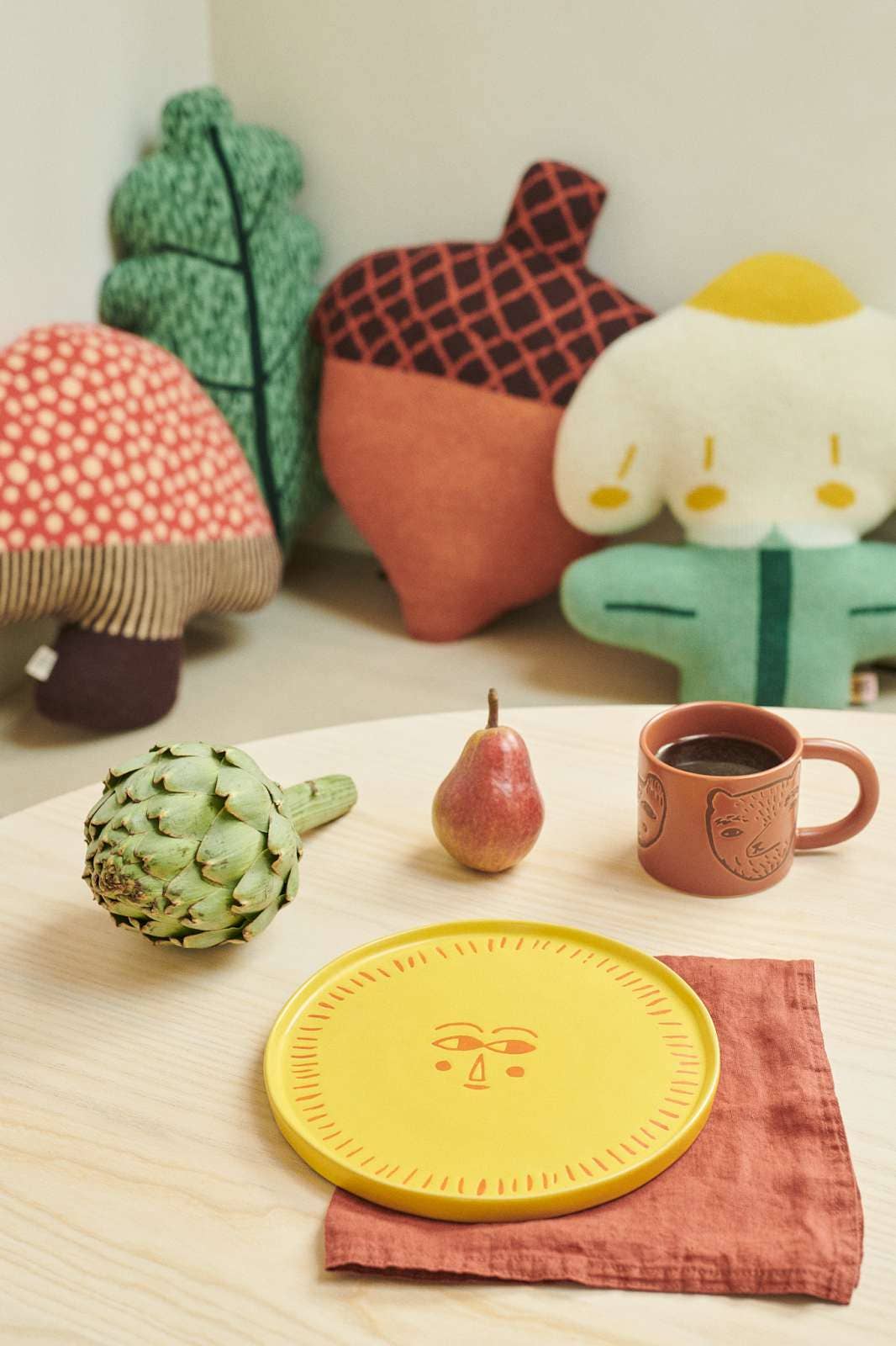 A styled tabletop scene with a yellow plate featuring a simple face design, placed on a rust-colored napkin. A small terracotta mug with a face illustration sits beside it, along with a green artichoke, a pear, and a rust-colored napkin.