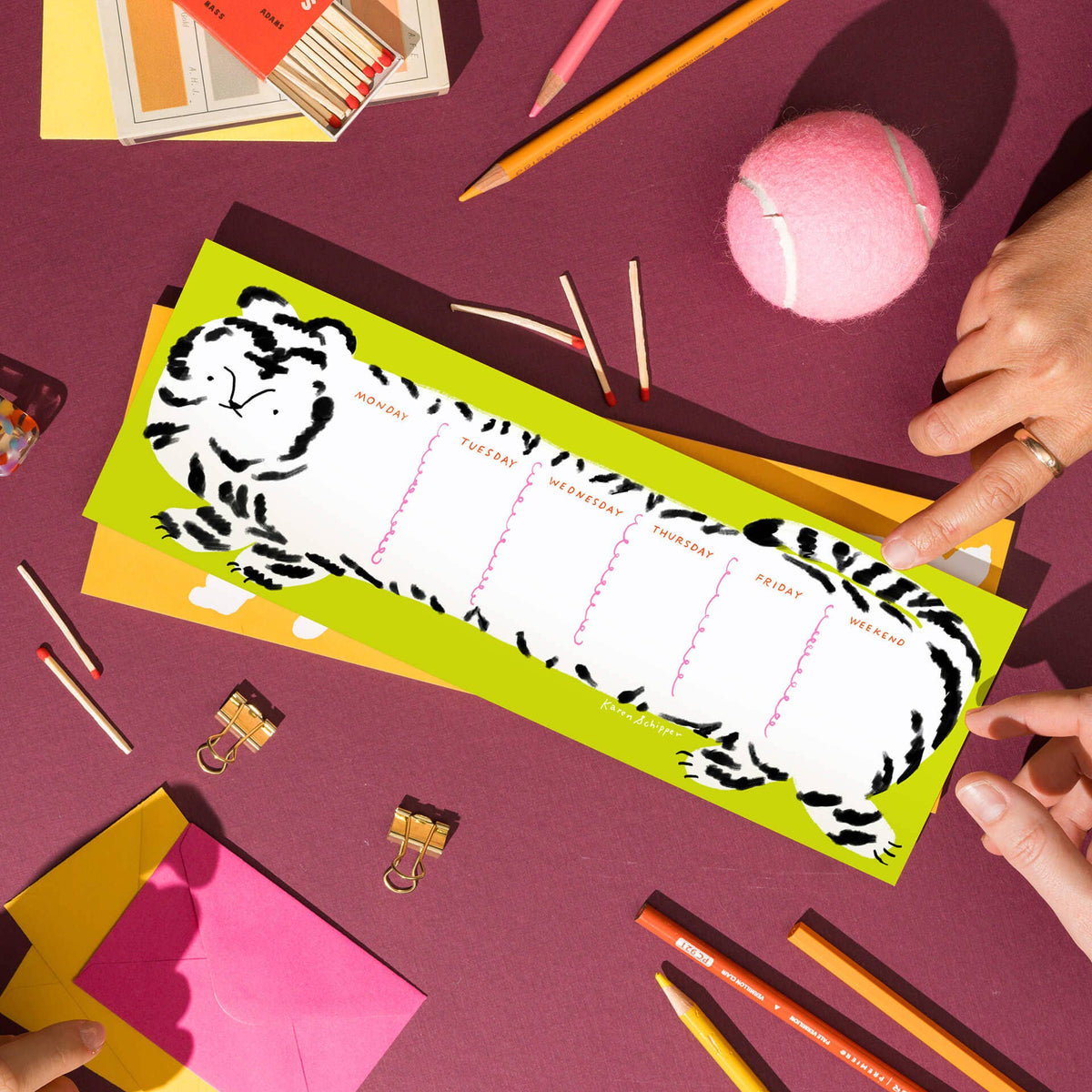 A colorful desk flat-lay featuring a long, bright lime green tiger-striped notepad with white writing sections. The notepad is surrounded by various desk accessories including pink sticky notes, yellow and orange pencils, binder clips, a pink tennis ball, and a matchbox labeled 'LOUSIERS'.