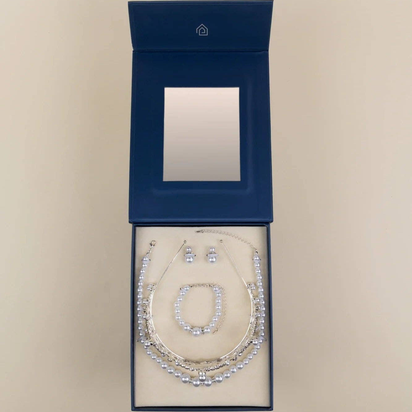 A jewelry set displayed inside an open dark blue presentation box with a mirror on the inside lid. The set includes a silver-toned necklace, bracelet, and earrings with clear rhinestone-like stones, arranged on a cream insert against a light beige background.