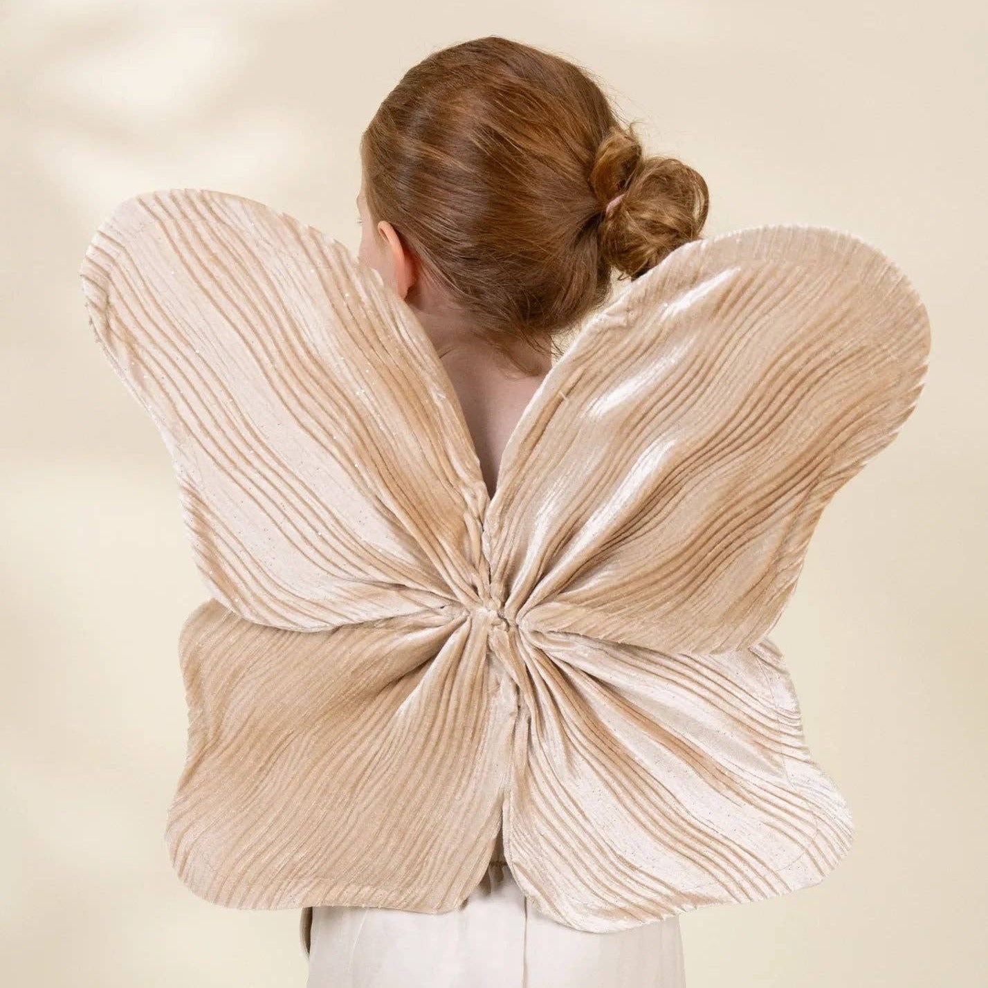 A child viewed from behind wearing a large pleated butterfly-wing costume accessory in a warm beige or cream tone, against a soft cream studio background.