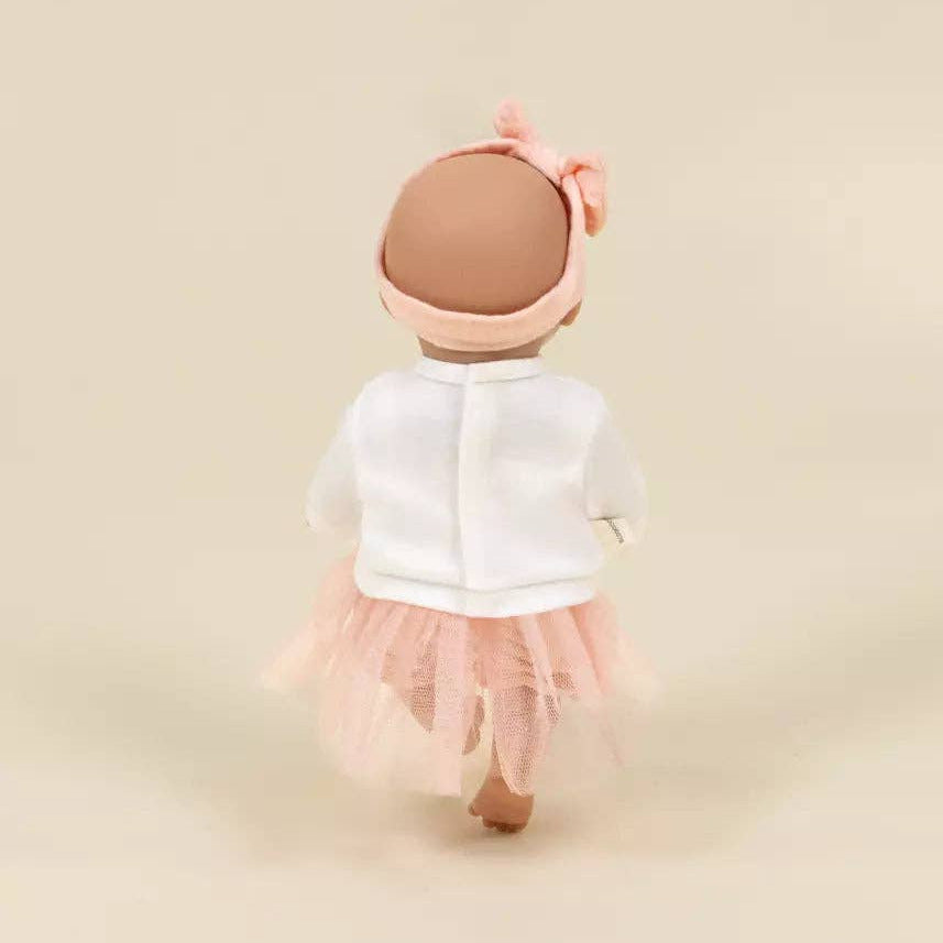 A doll viewed from behind wearing a white long-sleeve top, a light pink tutu skirt, and a matching pink bow headband, set against a solid beige background.