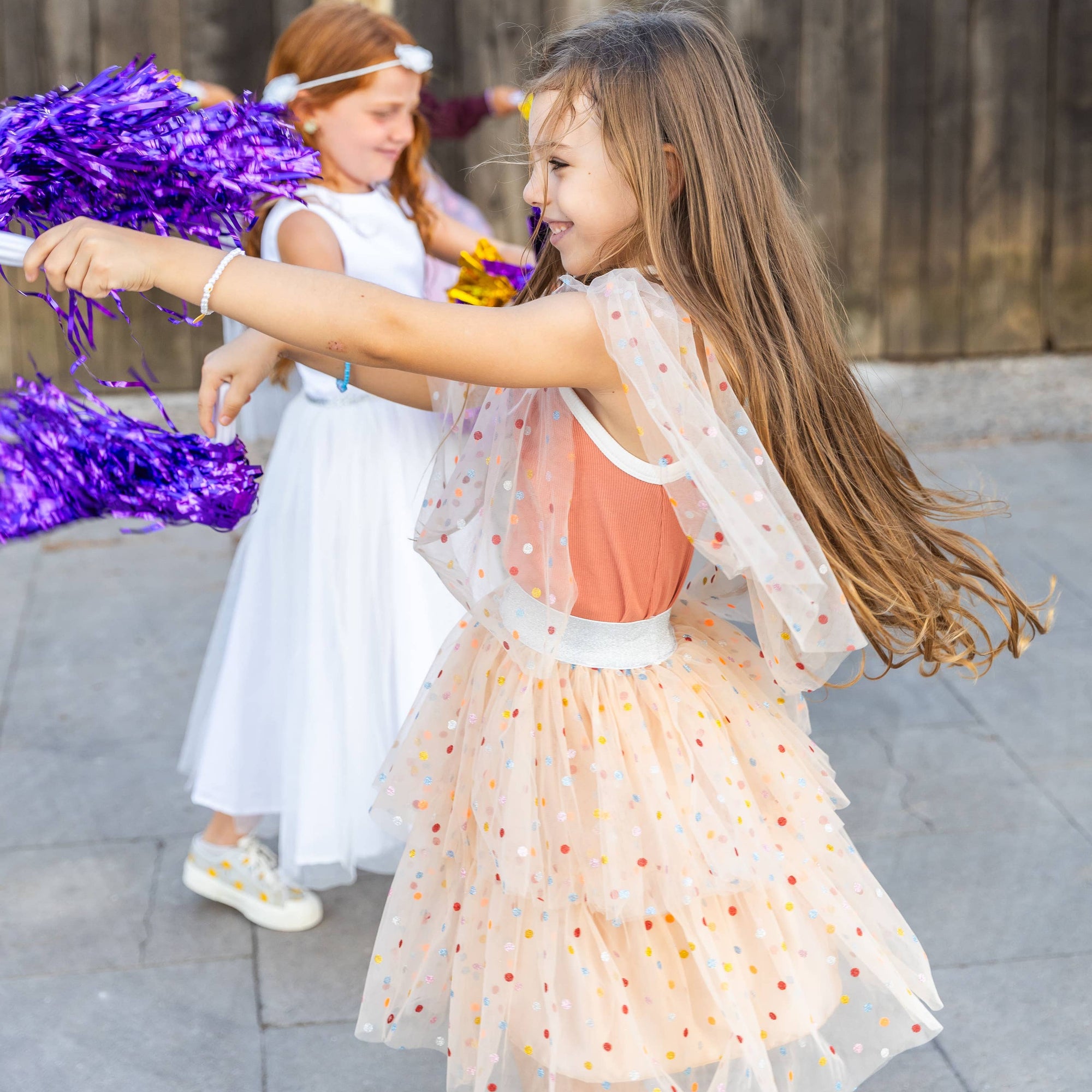 Two young girls in dresses with pom-poms on a stone pavement.