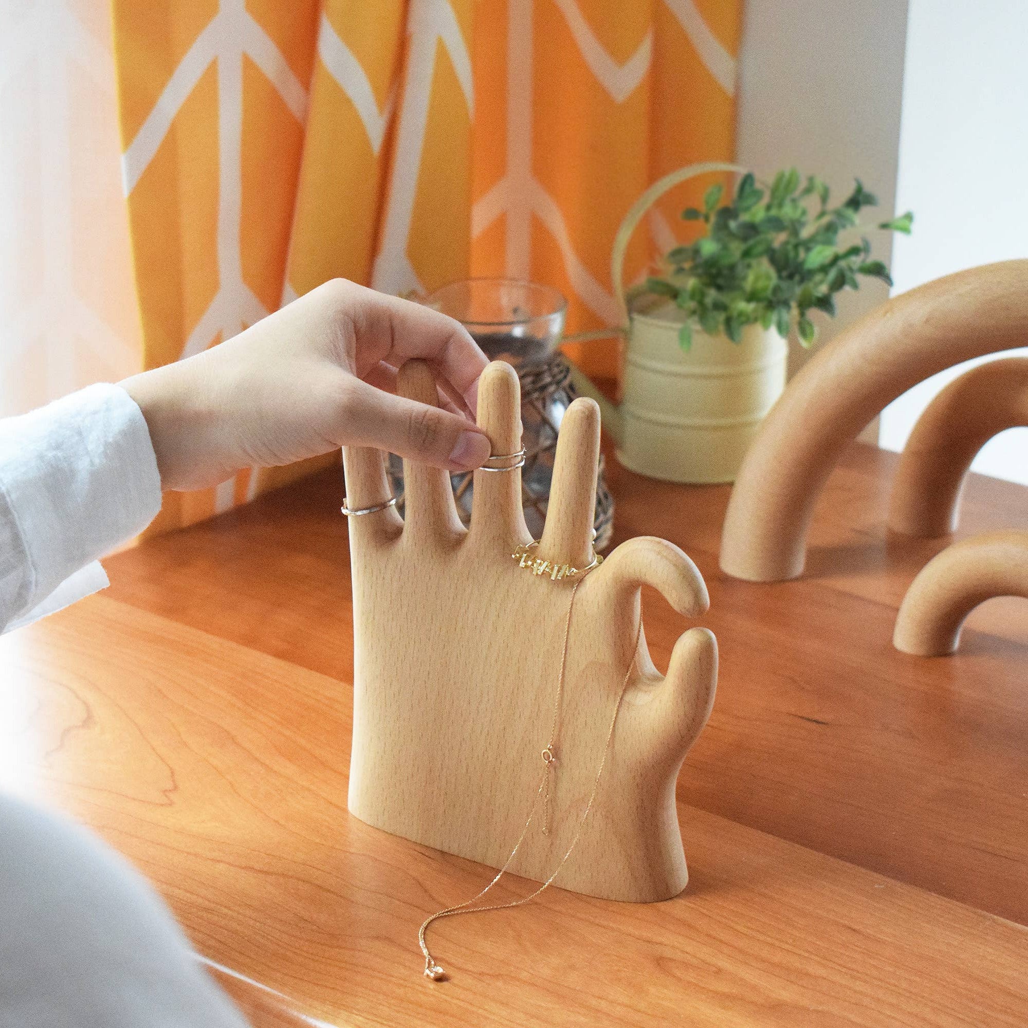 Person holding a ring above a wooden hand-shaped jewelry holder on a wooden table.