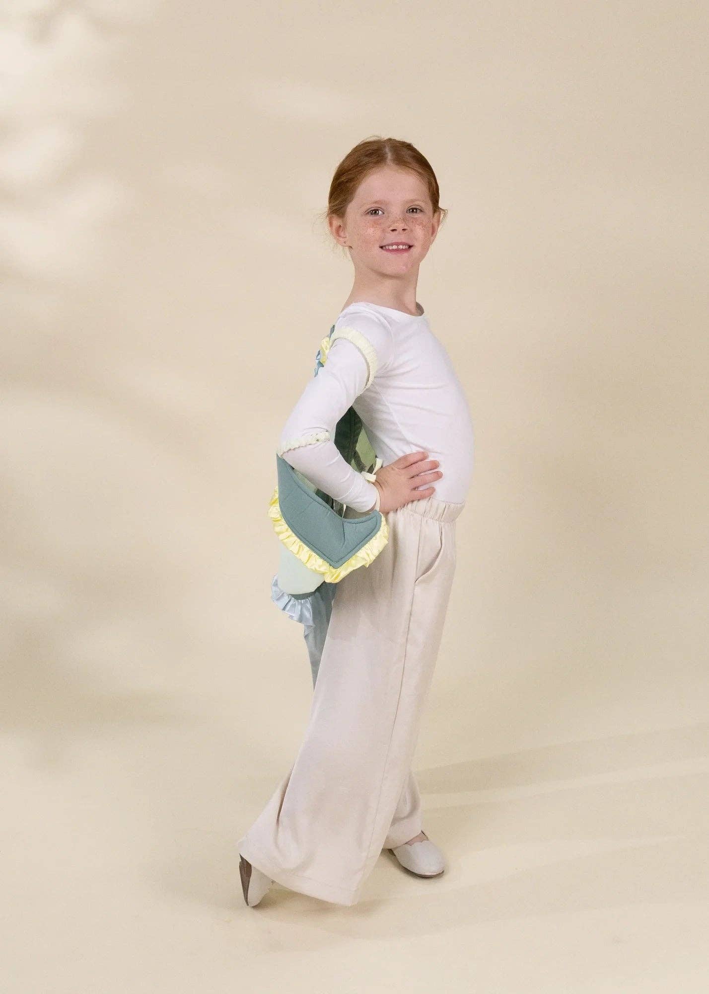 A young girl with red hair stands in a studio wearing a white long-sleeve top, cream wide-leg pants, and gray shoes, while holding a mint green and white handbag with yellow trim.