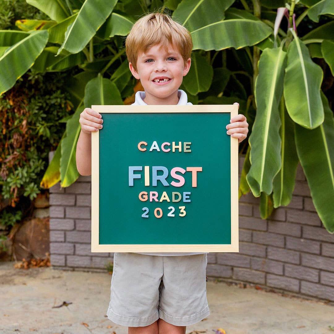Child holding a sign with 'CACHE FIRST GRADE 2023' in front of green foliage.