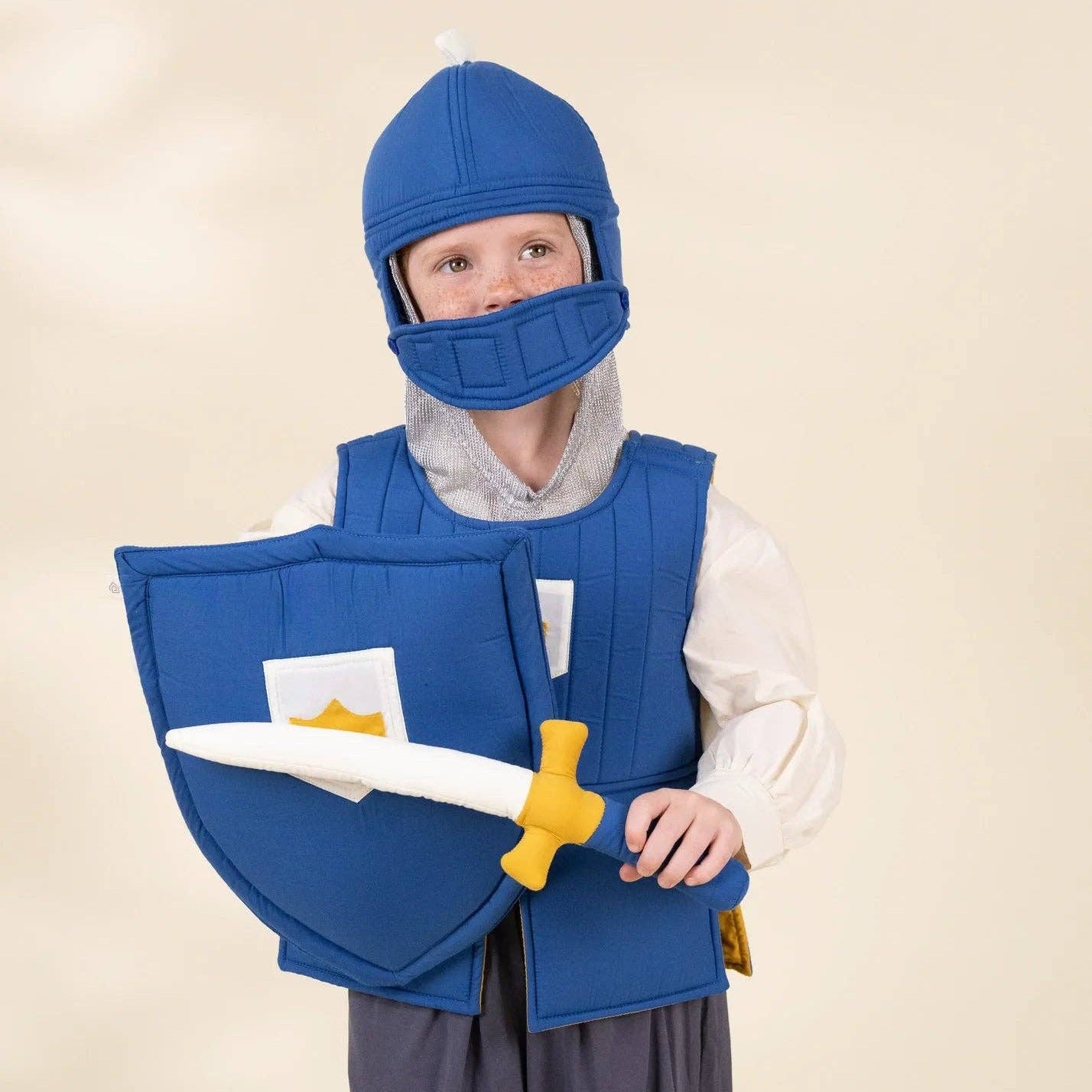 A child wearing a medieval knight costume, including a blue padded helmet, blue breastplate, white tunic, and gray pants, holding a blue shield and toy sword against a plain beige studio background.