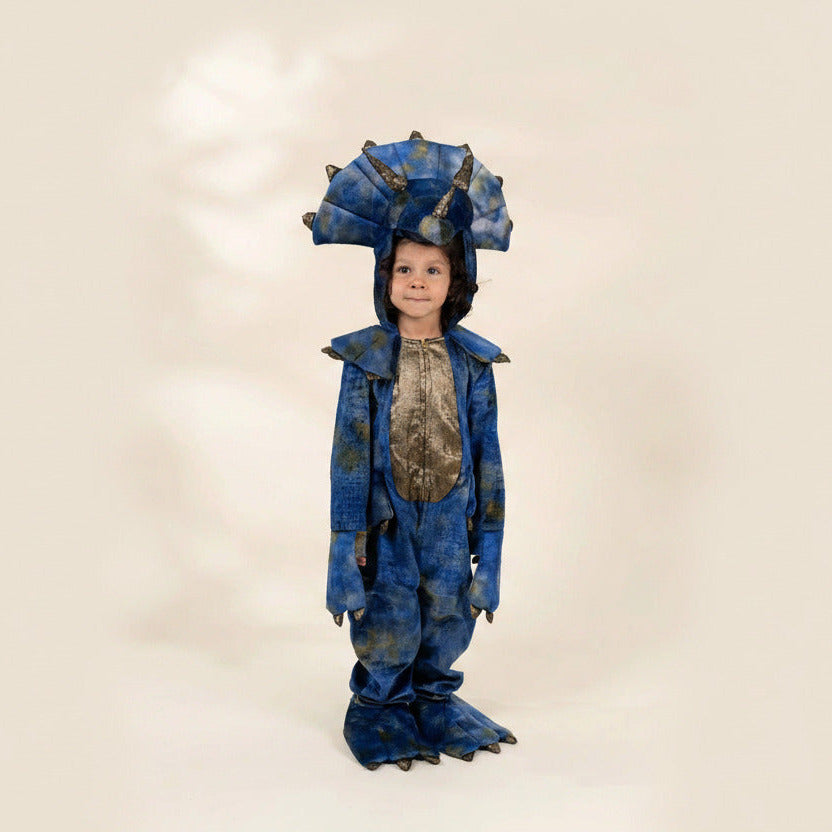 A child wearing a blue dinosaur costume with a large horned headpiece, textured blue jumpsuit, and clawed hands and feet, standing against a plain beige studio background.