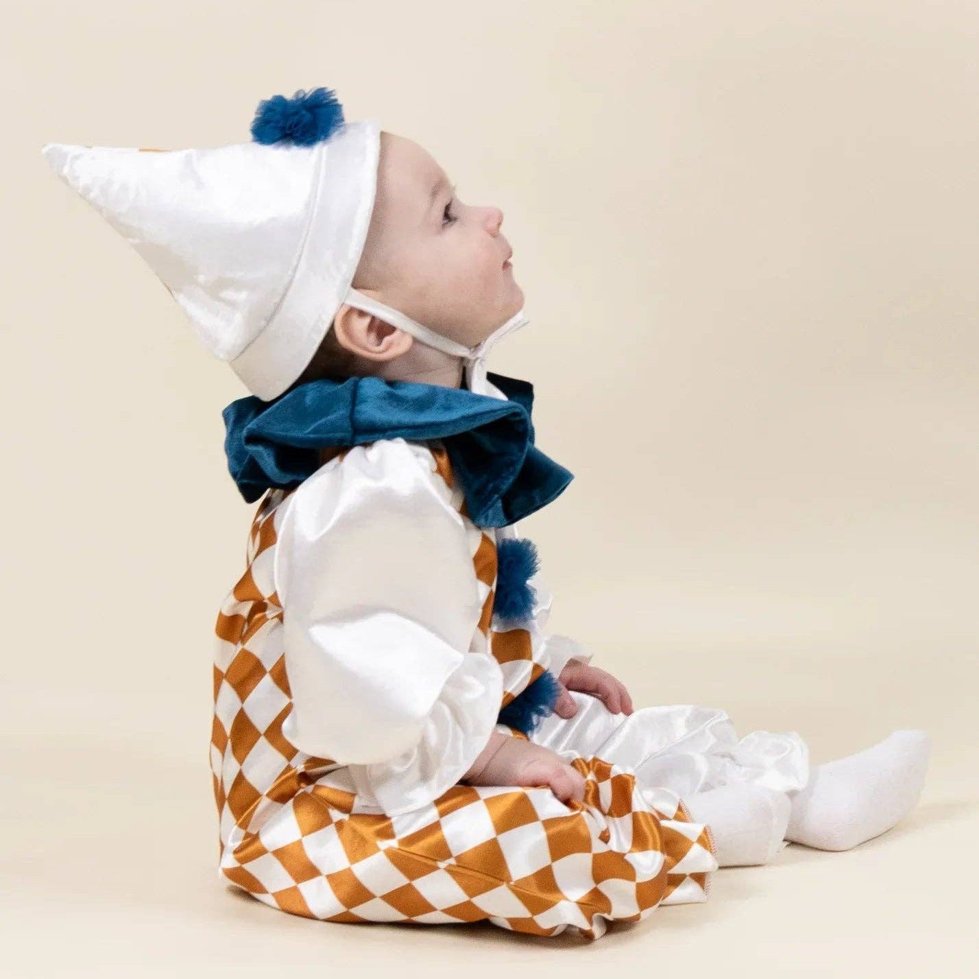 A baby wearing a white and orange checkered costume with a white pointed hat, blue pom-pom, and a dark blue ruffled collar, sitting against a plain beige background.