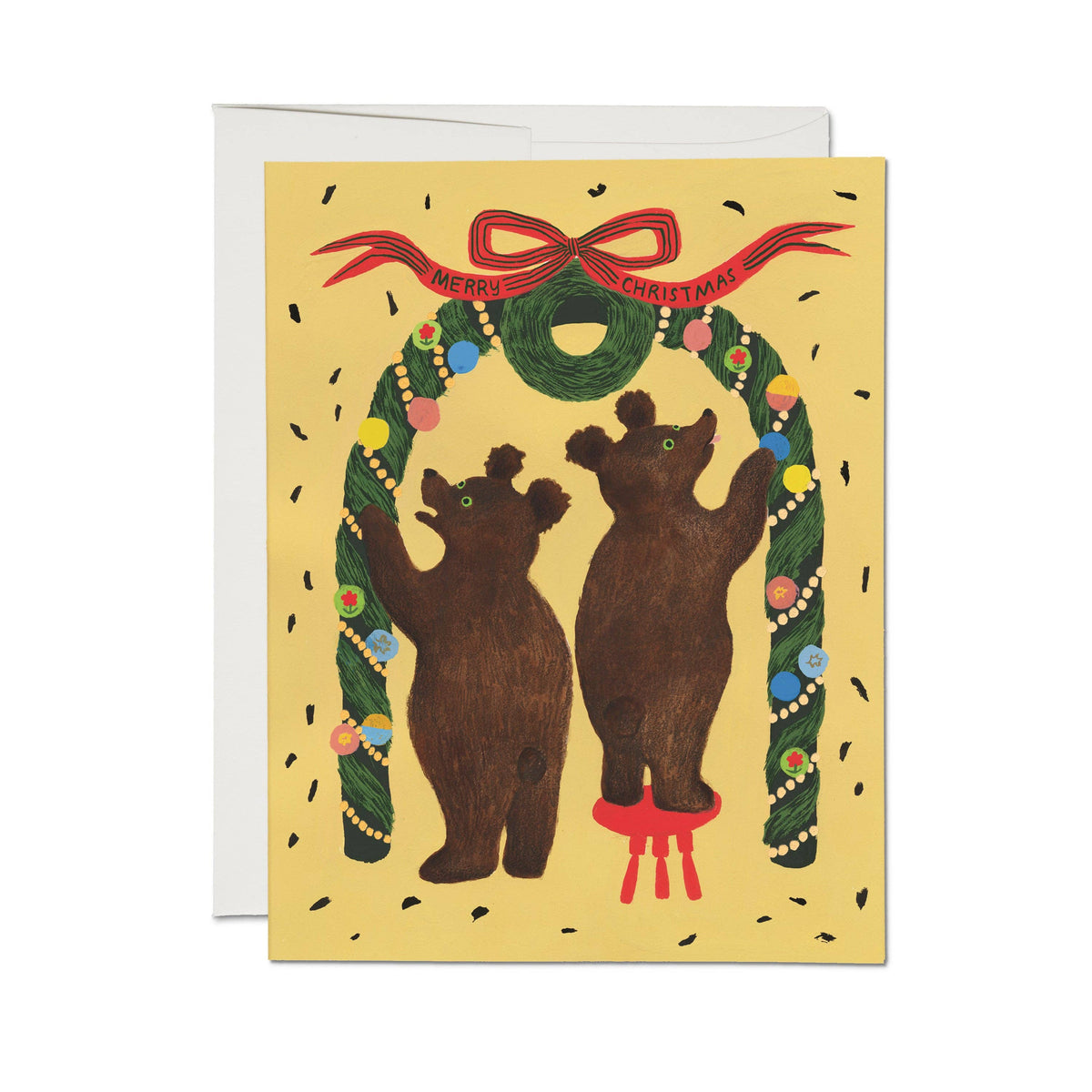 A Christmas greeting card featuring two brown bears standing under a green wreath decorated with colorful ornaments and a red bow. The card has a pale yellow background with scattered black confetti, and a white envelope is partially visible behind it.