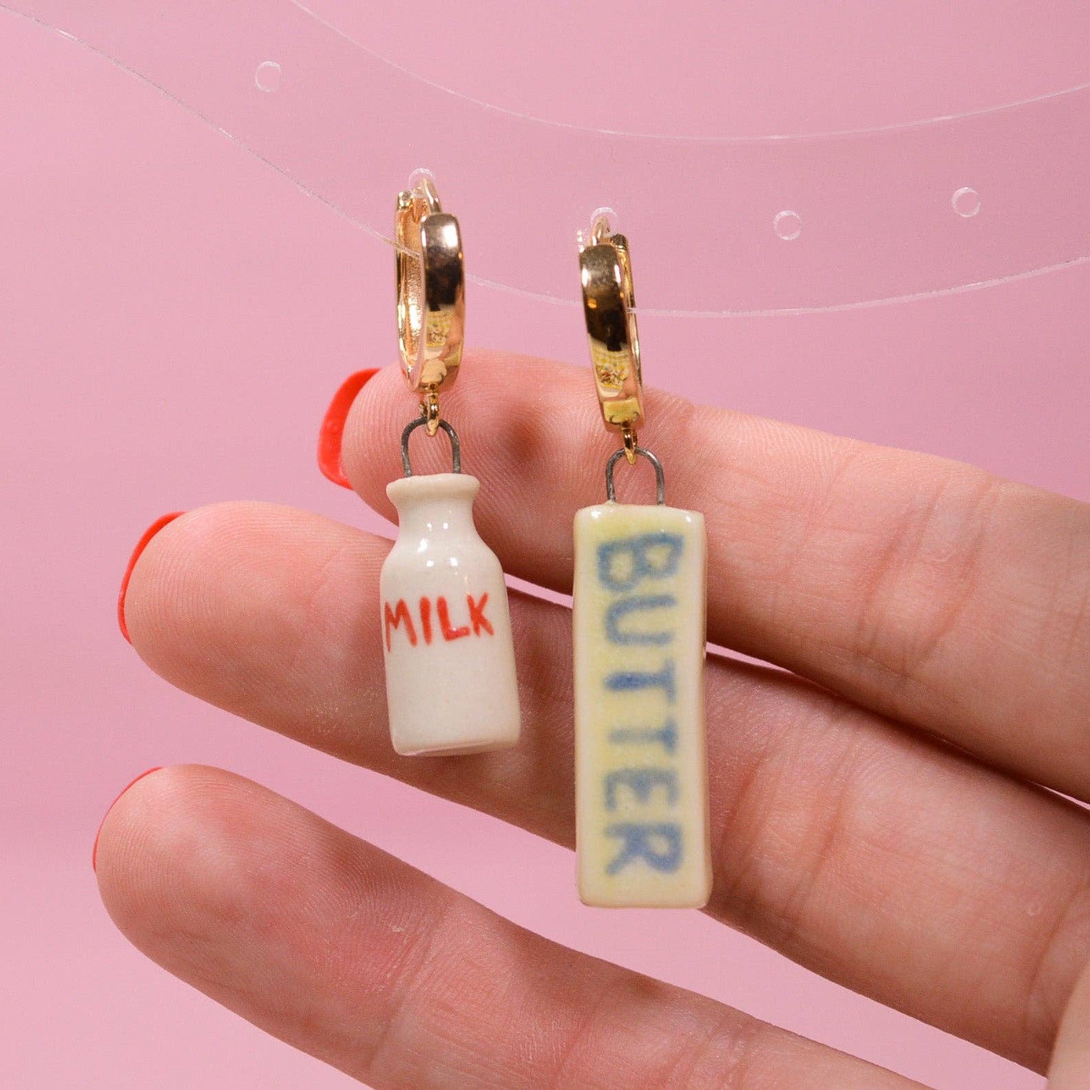 Milk bottle and butter bar earrings held between fingers against a pink background