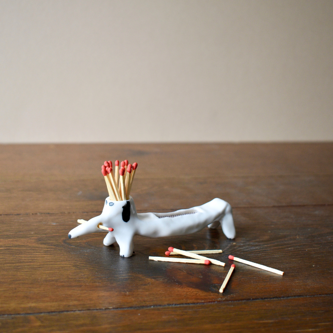 A white porcelain dog figurine with matchsticks inserted into its tail, placed on a wooden surface.