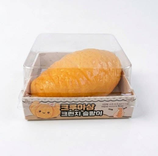 A packaged yellow-orange novelty food item shaped like a banana or bread loaf, displayed inside a clear plastic clamshell box with a printed cardboard base featuring Korean text and a cartoon illustration.