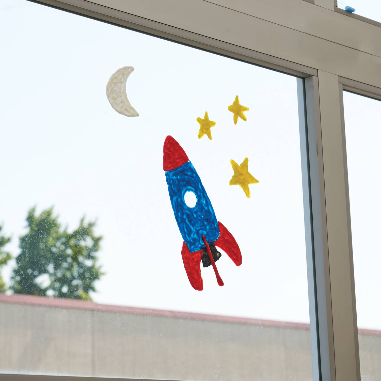A colorful rocket ship window sticker with a blue body, red nose and fins, yellow stars, and a gray crescent moon, applied to a glass window pane.