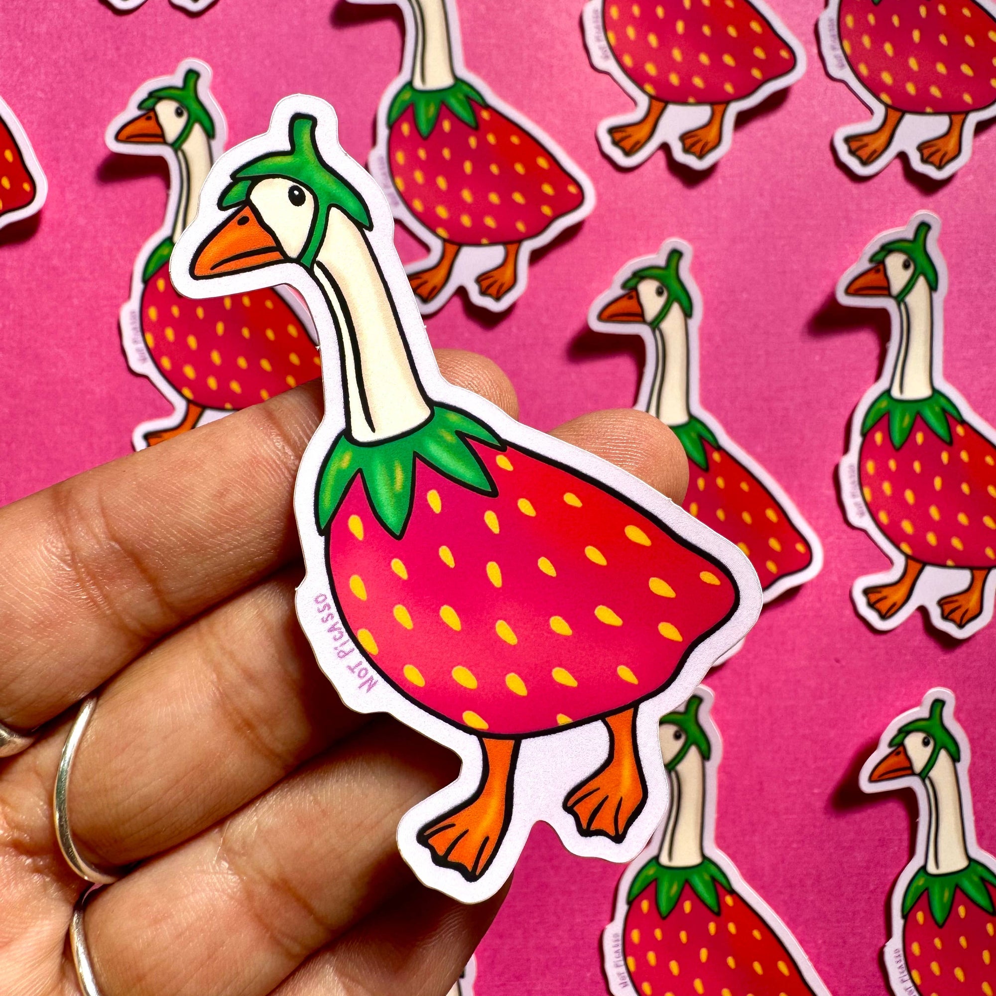 Hand holding a strawberry-themed duck sticker against a pink background with more stickers.
