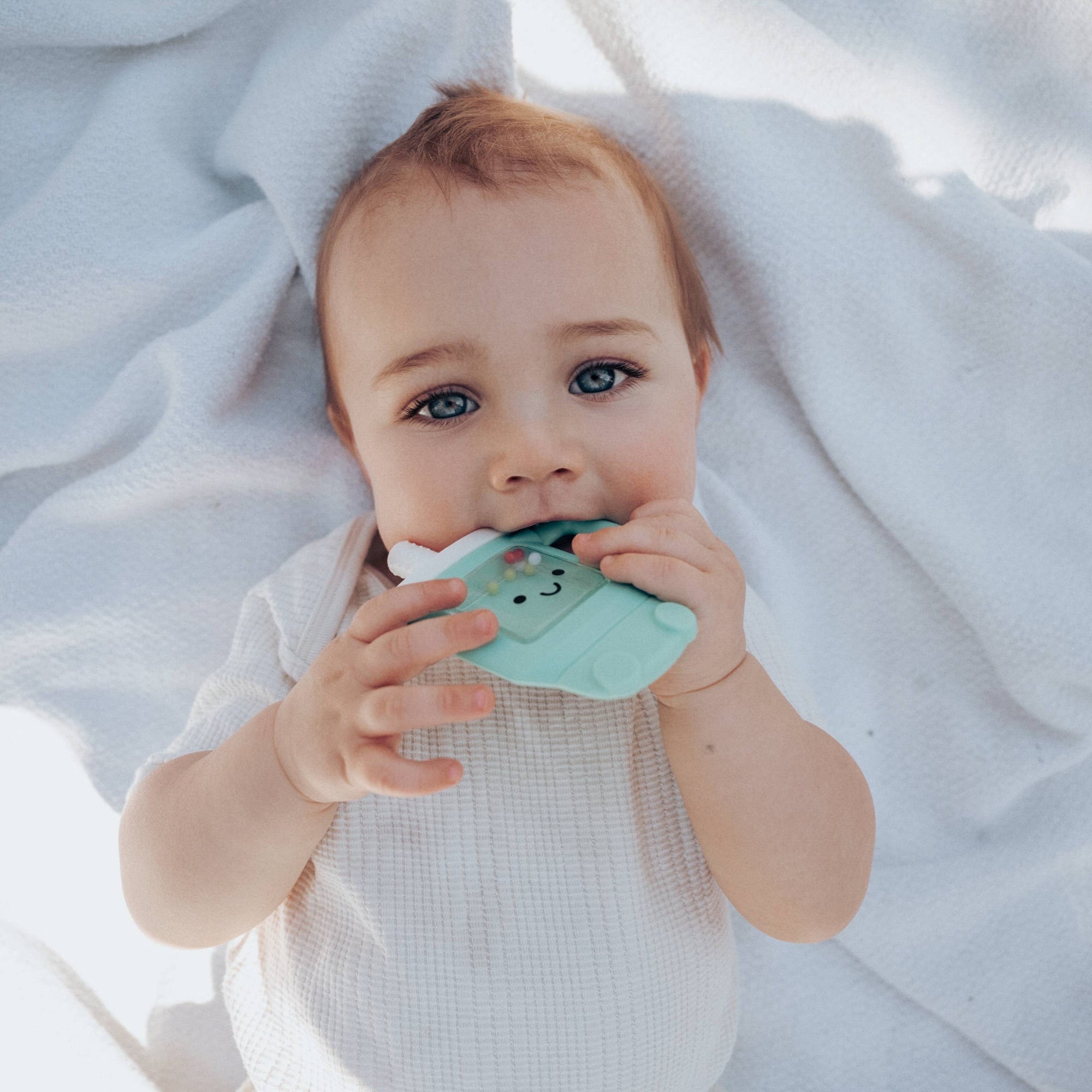 Baby holding a green teething toy on a white blanket