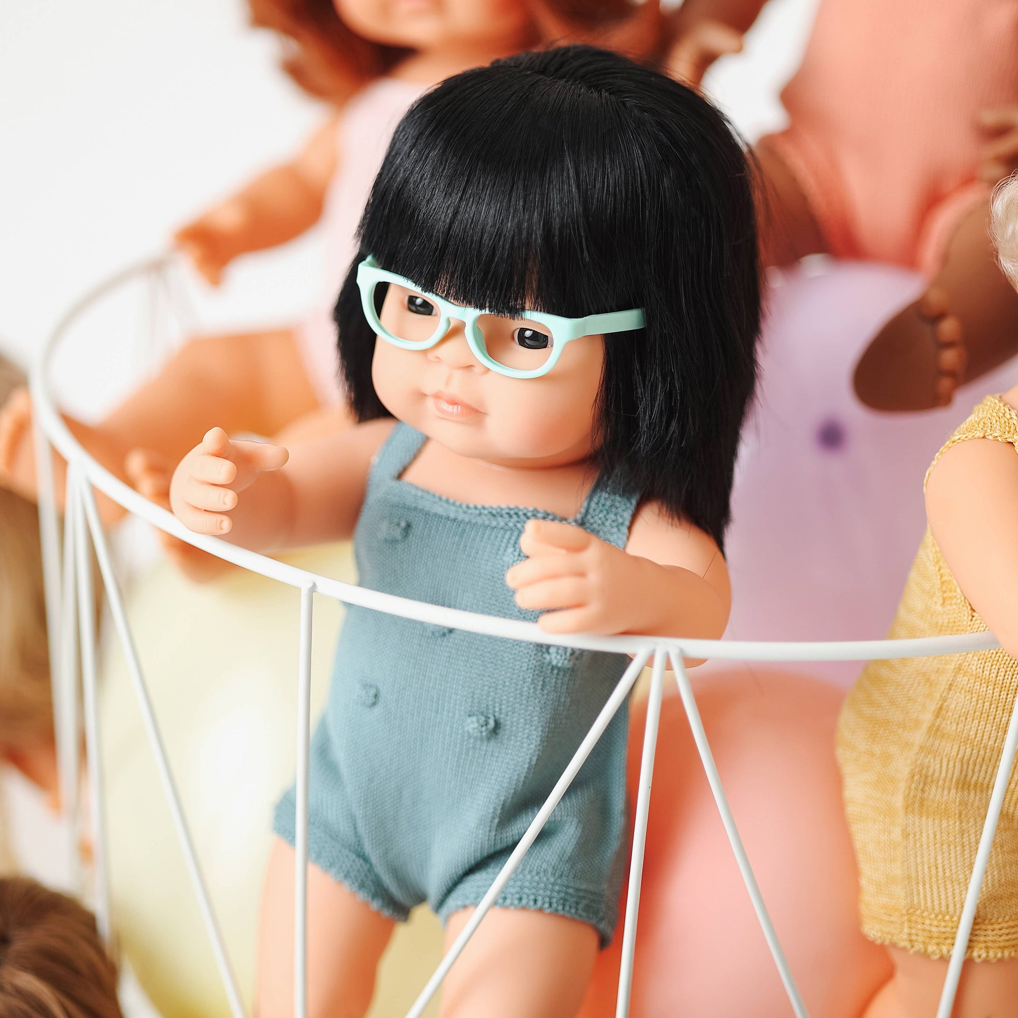 Doll with black hair and glasses sitting in a crib with other dolls in the background.
