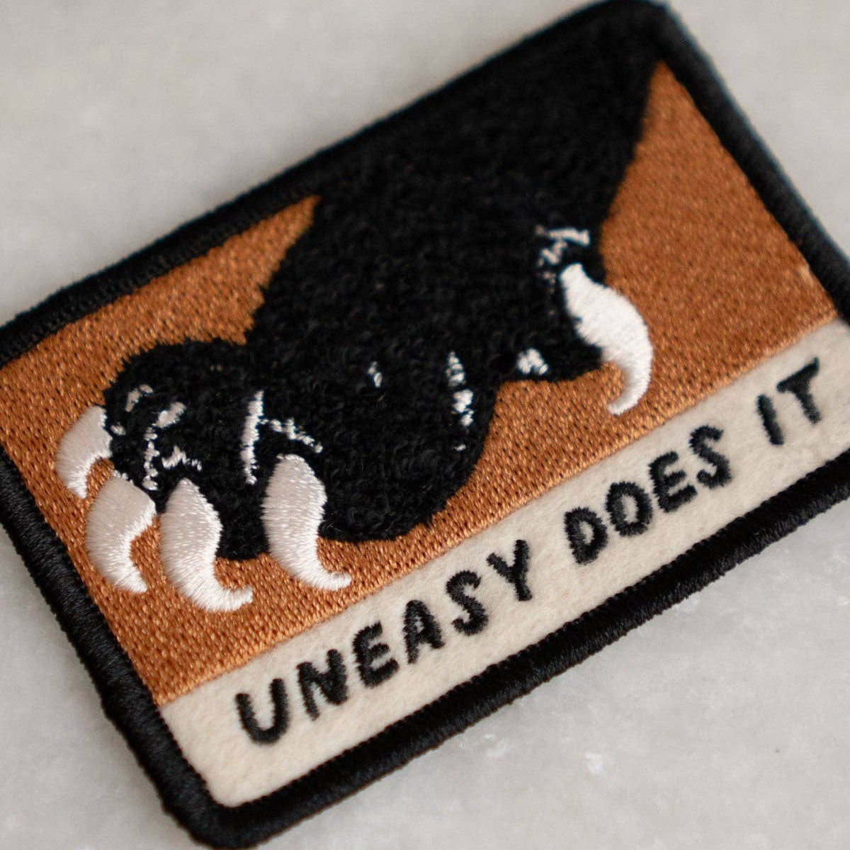 Embroidered patch with a claw design and 'Uneasy Does It' text on a textured surface.