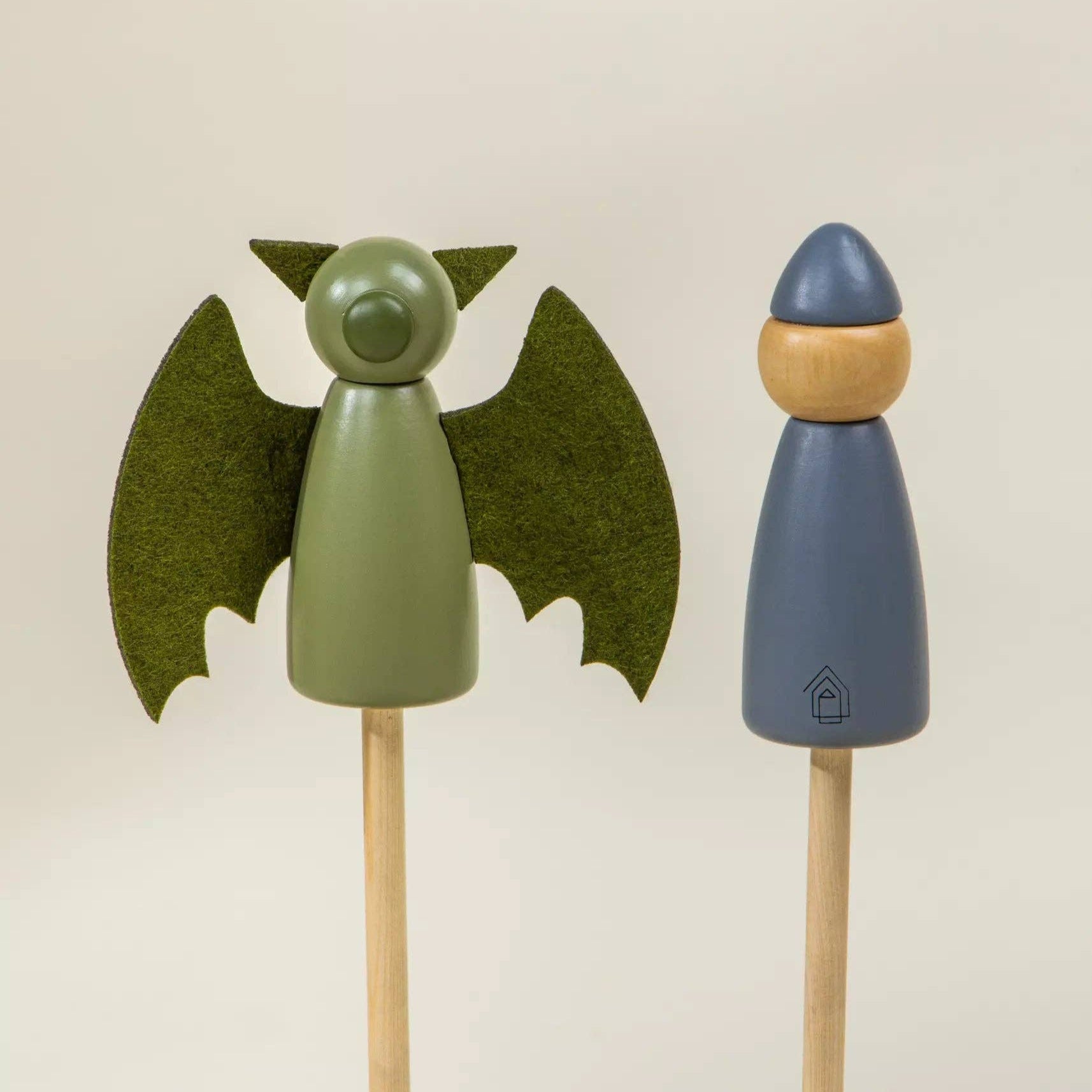 Two wooden toys, one green bat-like figure and one blue figure with a yellow hat, on a beige background.