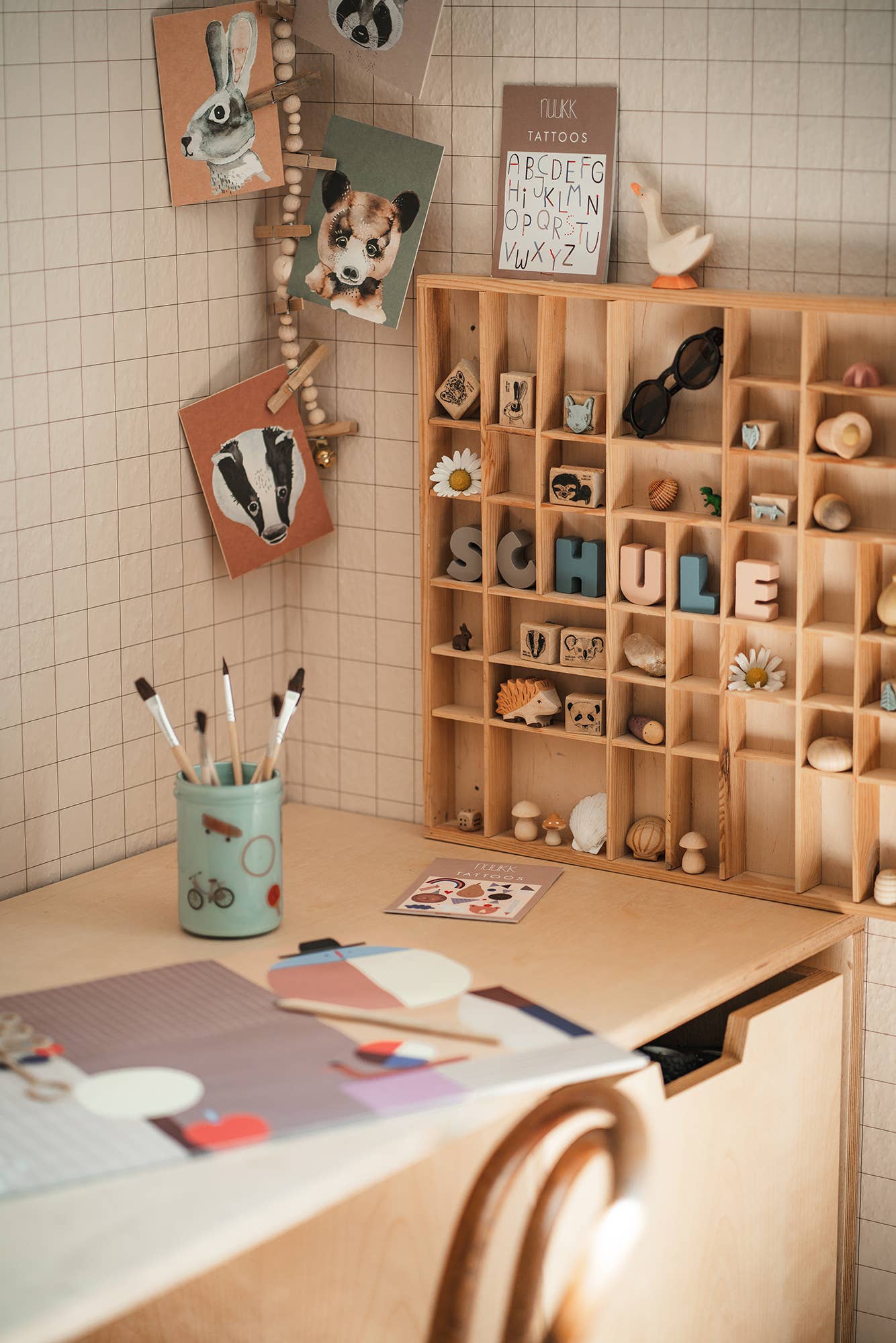 A cozy, warm-toned children's room corner featuring a light wood desk, a matching wooden toy organizer shelf filled with small educational and decorative items, and a tiled beige wall with whimsical hanging art cards.