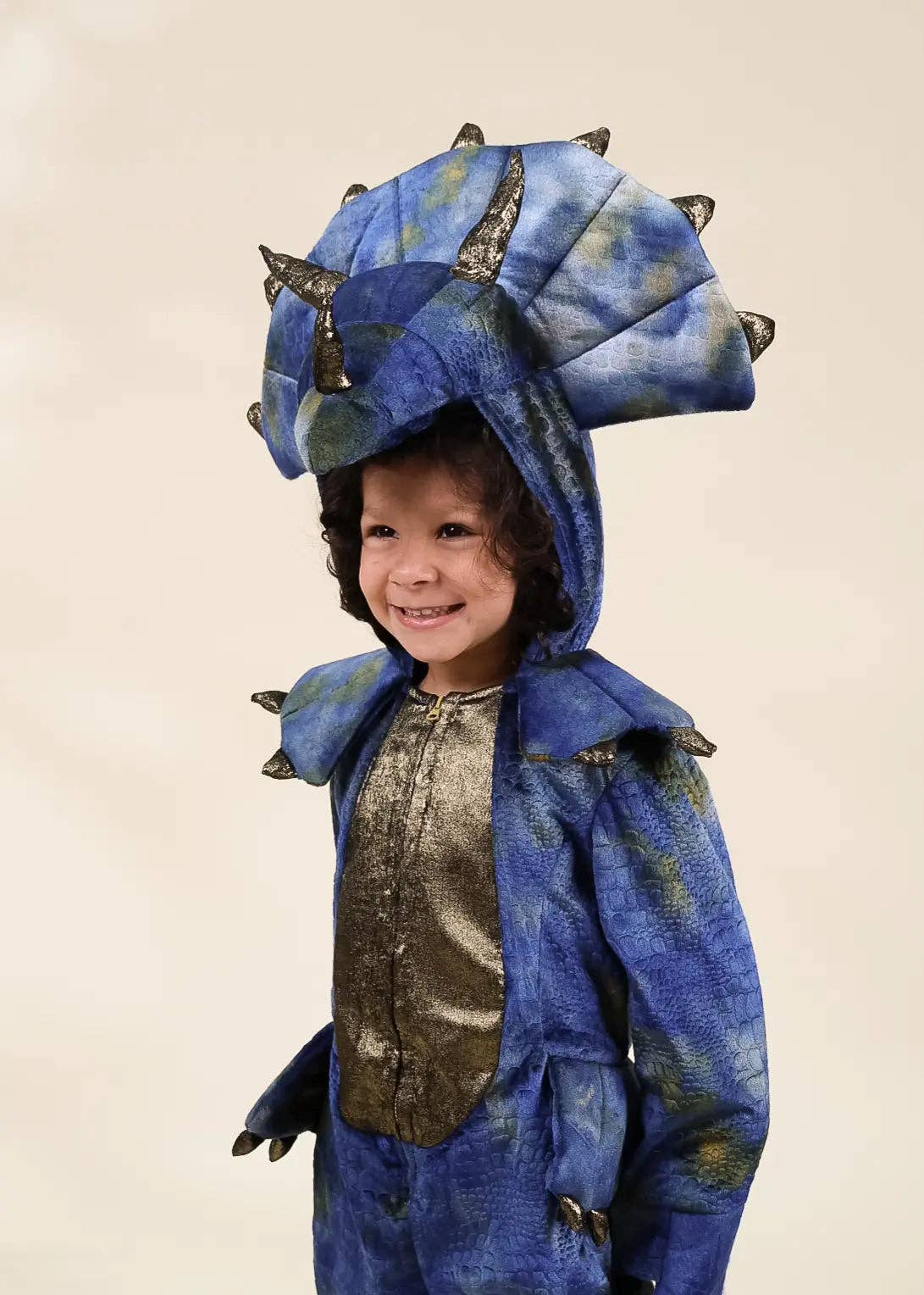 A young child wearing a blue dinosaur costume with a large matching hood featuring spikes, a textured blue body, and a metallic gold chest panel, photographed against a plain light beige background.