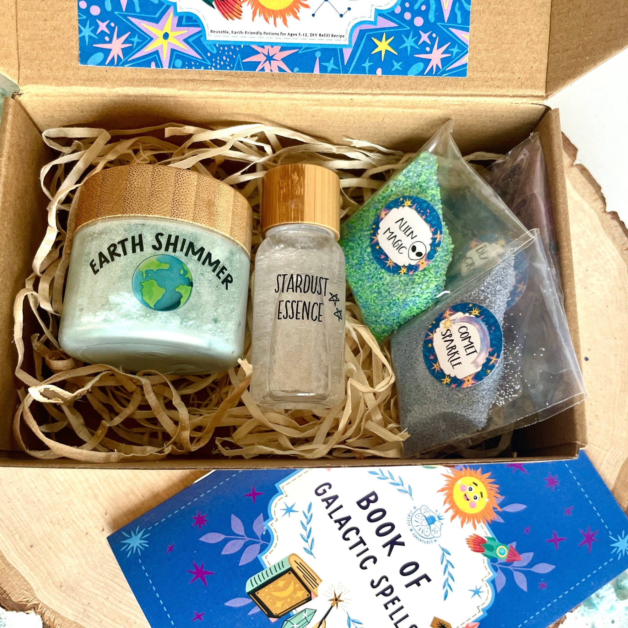 Box containing 'Earth Shimmer' candle, 'Starlight Essence' bottle, and other items with a 'Book of Galactic Spells' on a textured surface.