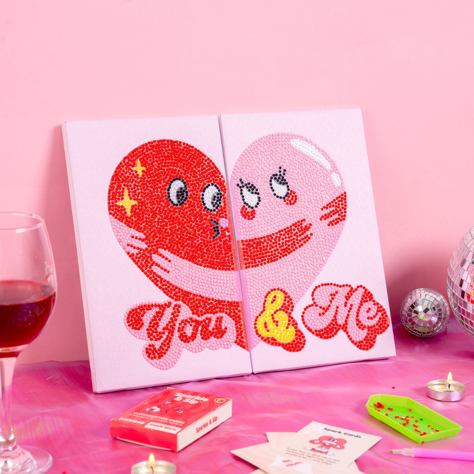 Two heart-shaped canvases with cartoon faces and 'You & Me' text on a pink background with a glass of red wine and disco ball.