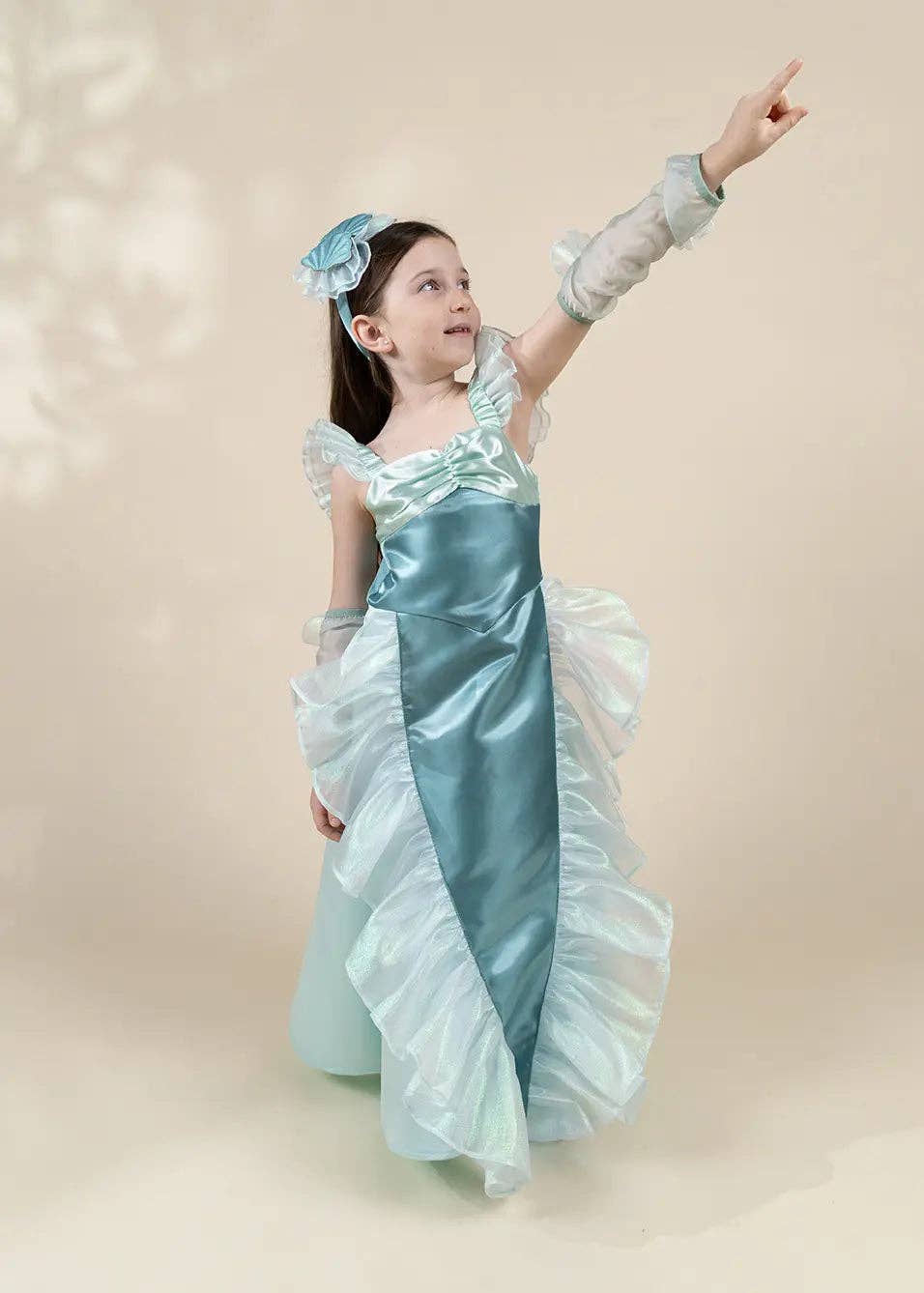 A young girl wearing a light blue and white princess-style dress with sheer long sleeves, a matching headband, and a pointed finger, standing against a warm beige studio background.