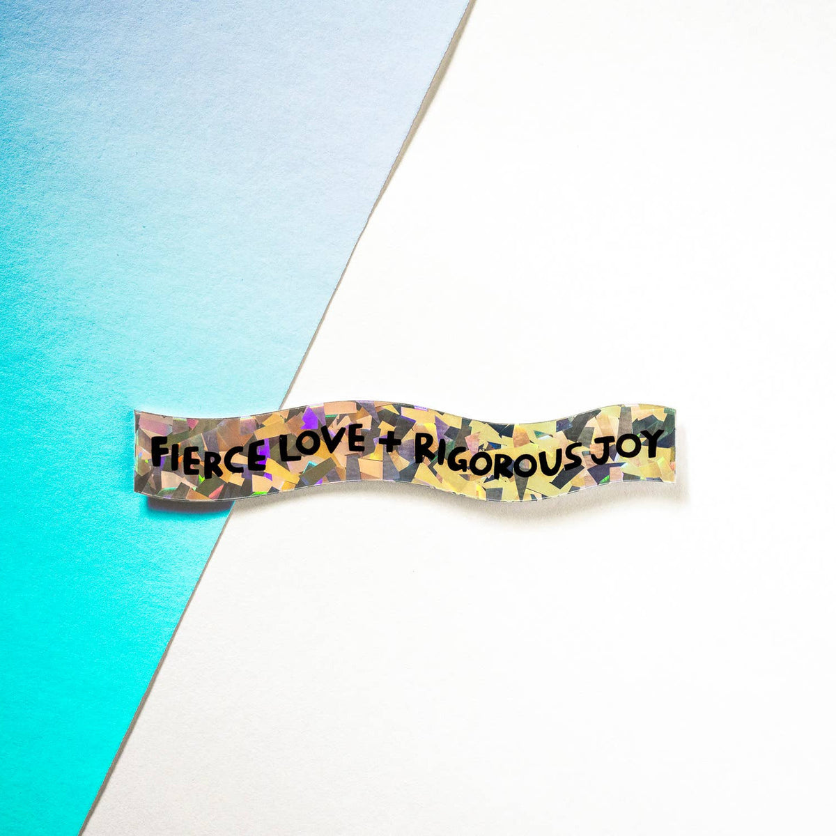 A small rectangular patch or sticker with the phrase “FIERCE LOVE + RIGOROUS JOY” in black text over a multicolor camouflage-like pattern, placed on a white surface with a diagonal turquoise-blue section behind it.