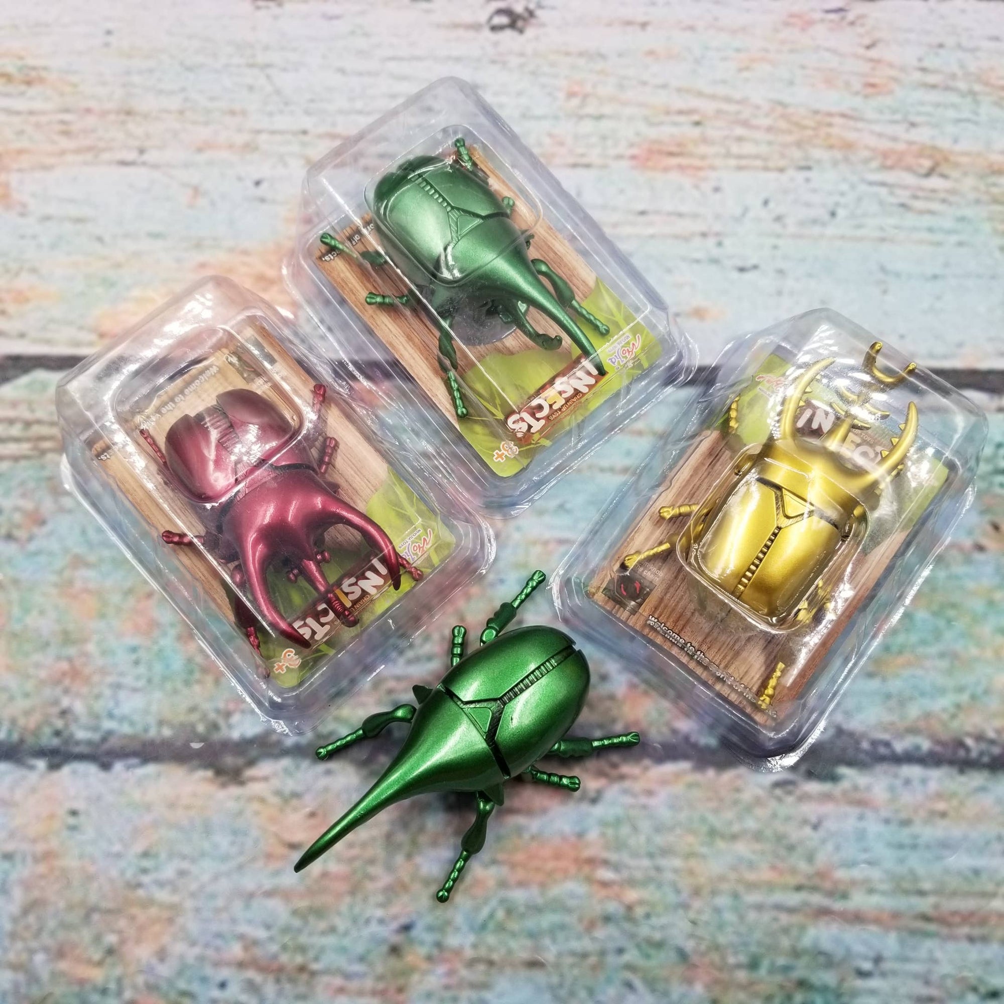 Three metallic beetle figurines in clear packaging on a textured surface