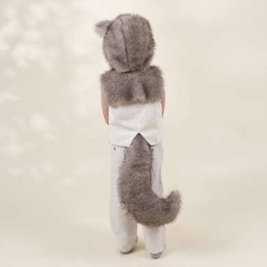 A child viewed from behind wearing a gray faux-fur hood with animal ears and a matching gray tail attached at the back, over a white top and white pants, against a soft cream studio background.