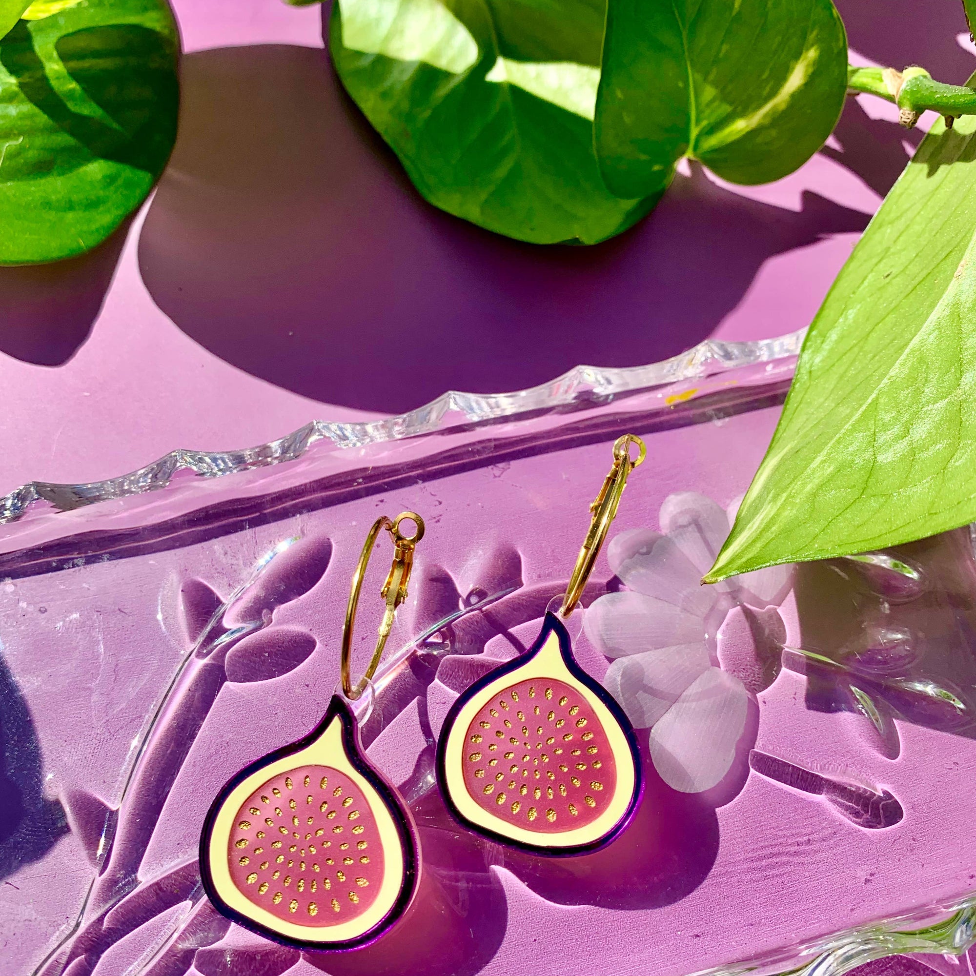A pair of earrings designed to resemble figs, with gold-colored hoops, displayed on a purple surface with green leaves in the background.