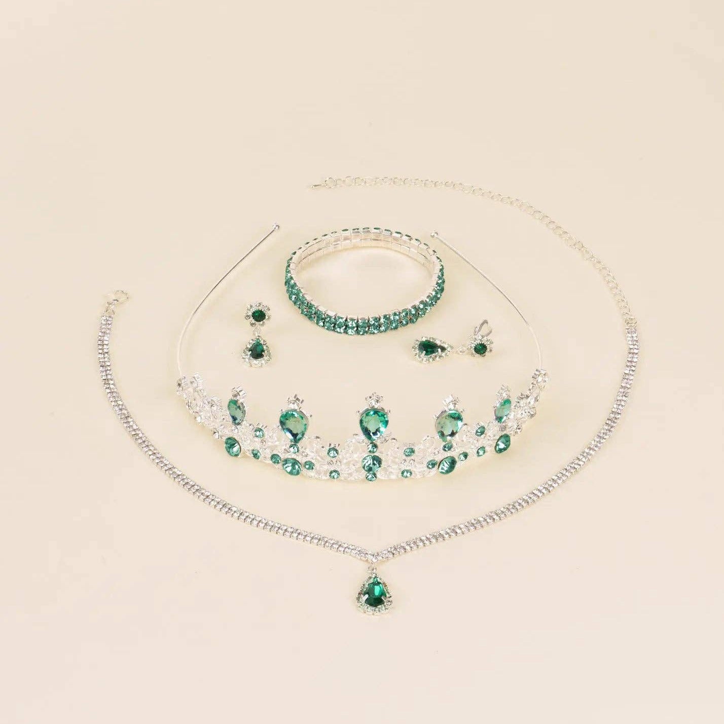 A jewelry set featuring a silver-tone necklace with a green teardrop pendant, a matching green beaded bracelet, a silver-tone tiara with green gemstones, and matching stud earrings, arranged on a light beige background.