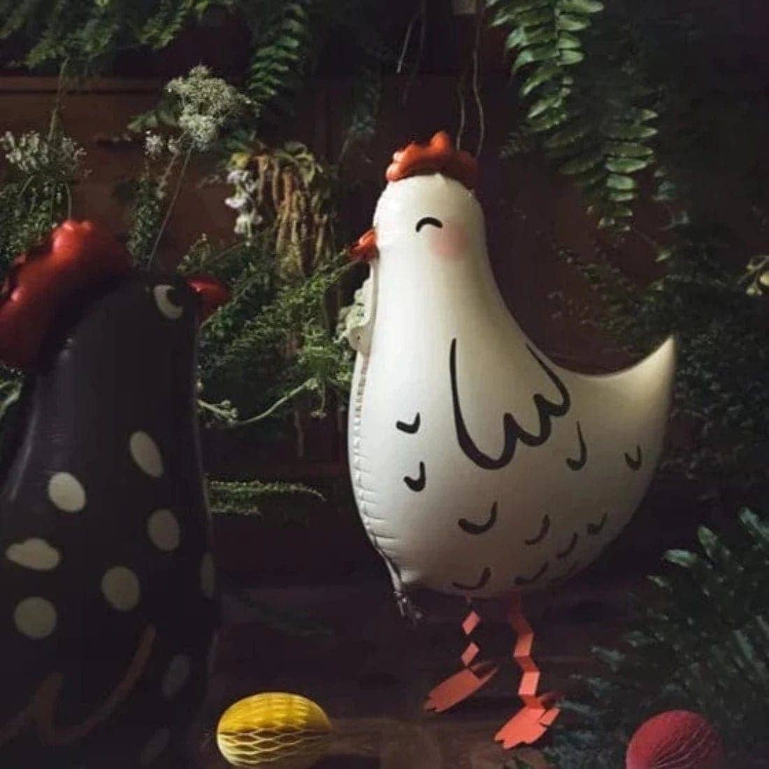 A white inflatable chicken decoration with black markings, orange comb and wattle, and orange ribbon-like legs, hanging among green foliage. Part of a black inflatable chicken with white polka dots is visible at the left edge.