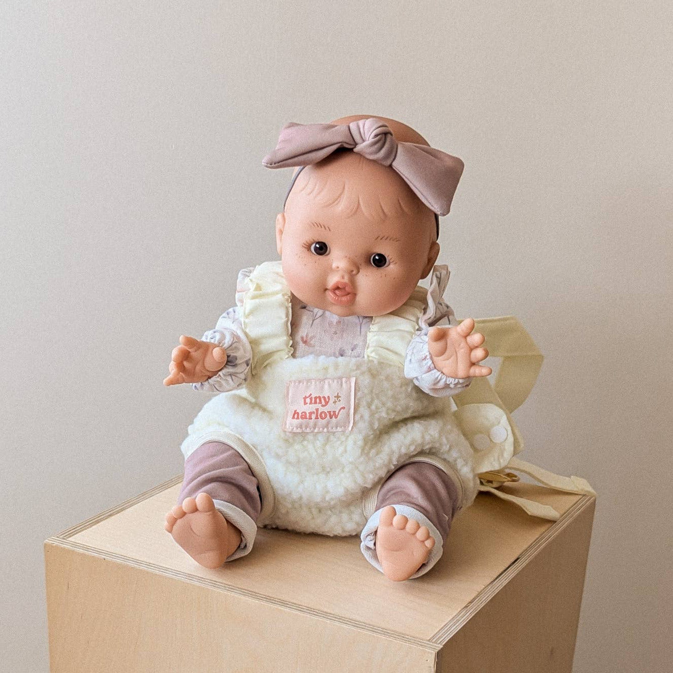 A realistic baby doll with a dusty pink bow headband and matching outfit, sitting on a light wood block against a plain beige wall.