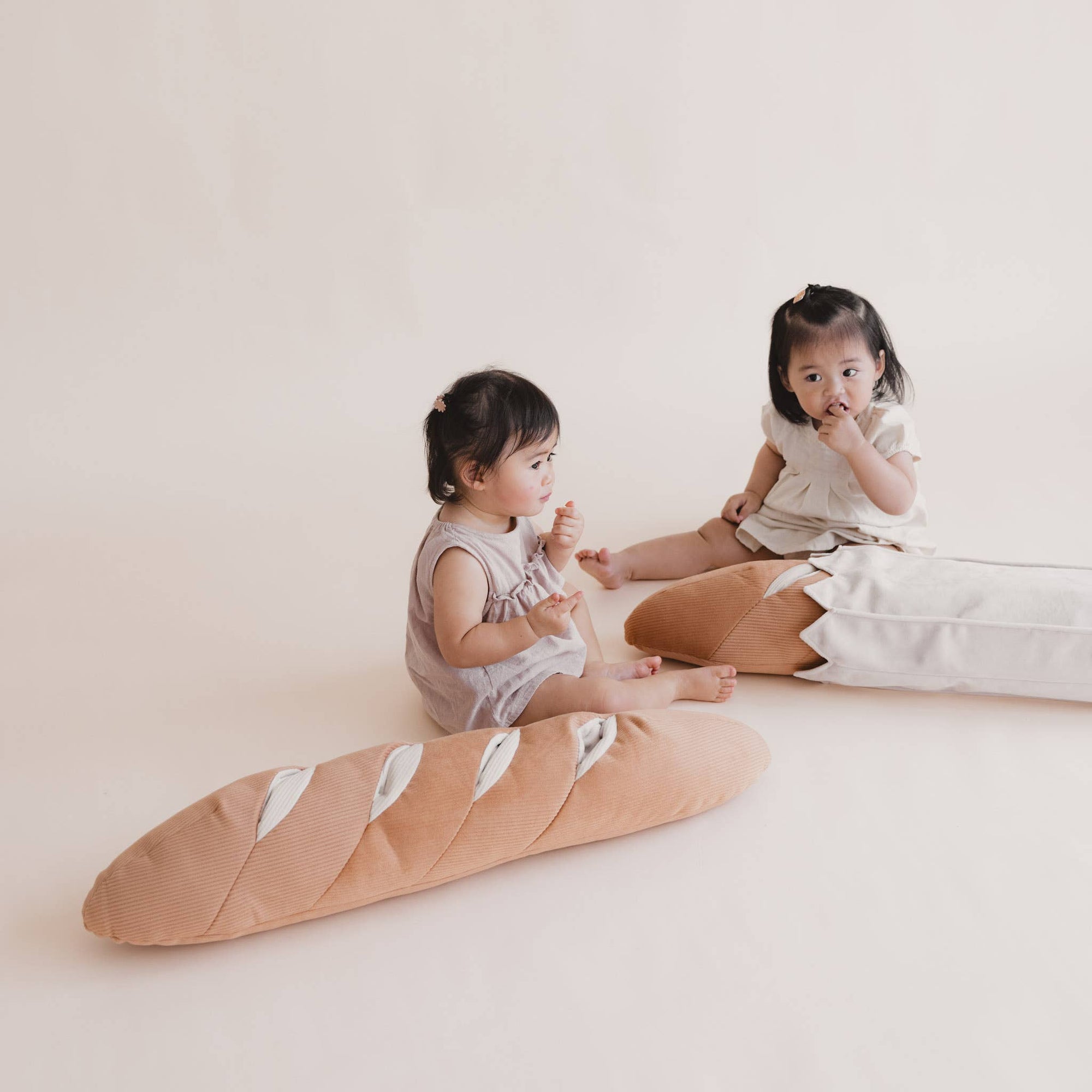 Two young children sitting on a large bread-shaped cushion against a plain background