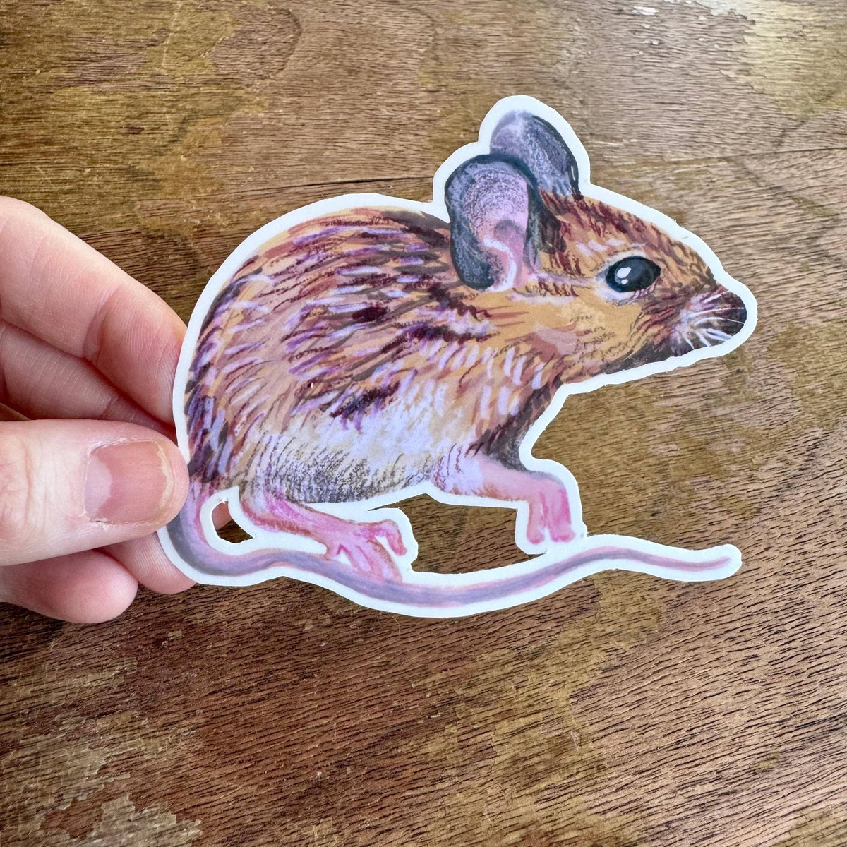 A hand holding a die-cut sticker of a brown and beige mouse with pink legs, shown against a wooden tabletop background.