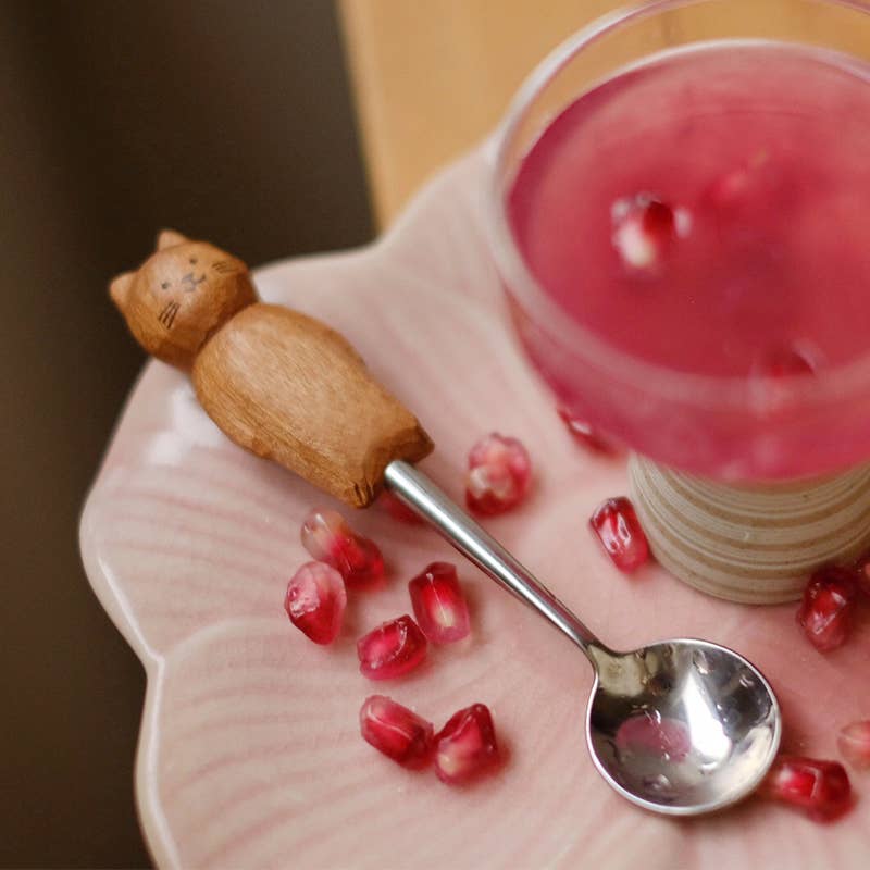 Wooden cat spoon, pomegranate seeds, and a spoon on a pink plate with a blurred background